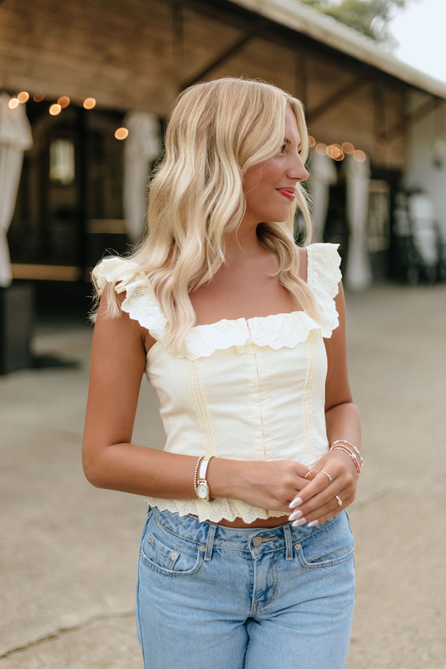 Woman in white ruffle crop top and light wash jeans poses casually against rustic building with string lights.
