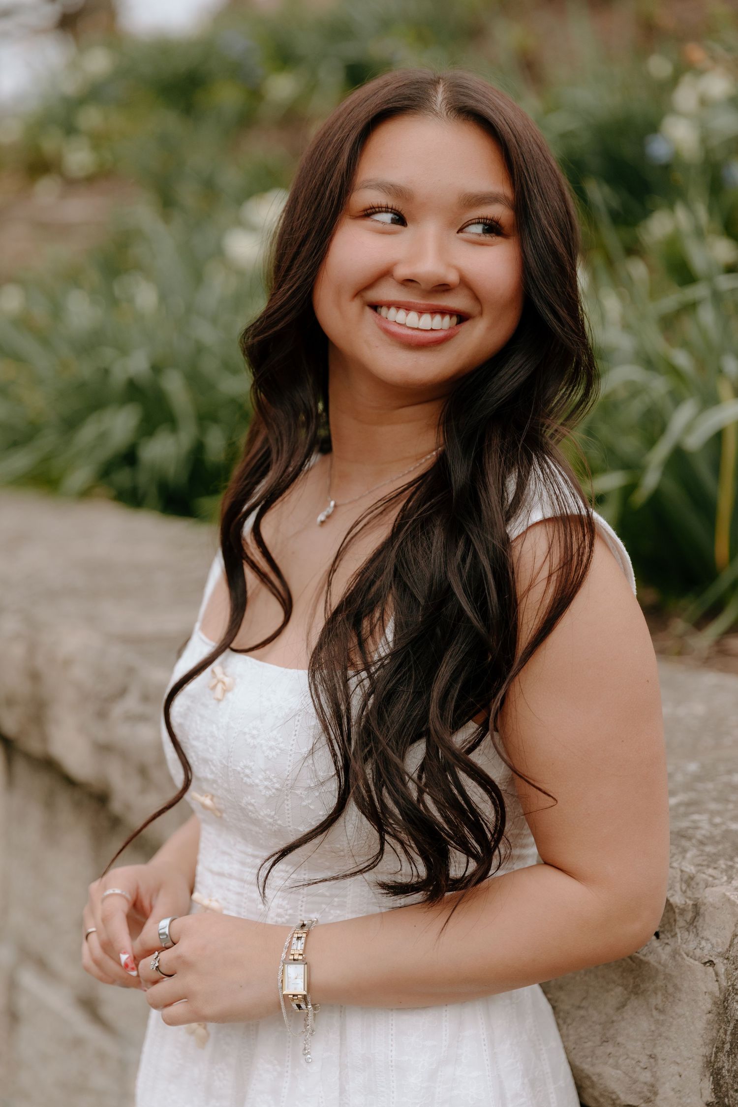 A woman in a white dress poses outdoors against a natural background with greenery and stone walls.
