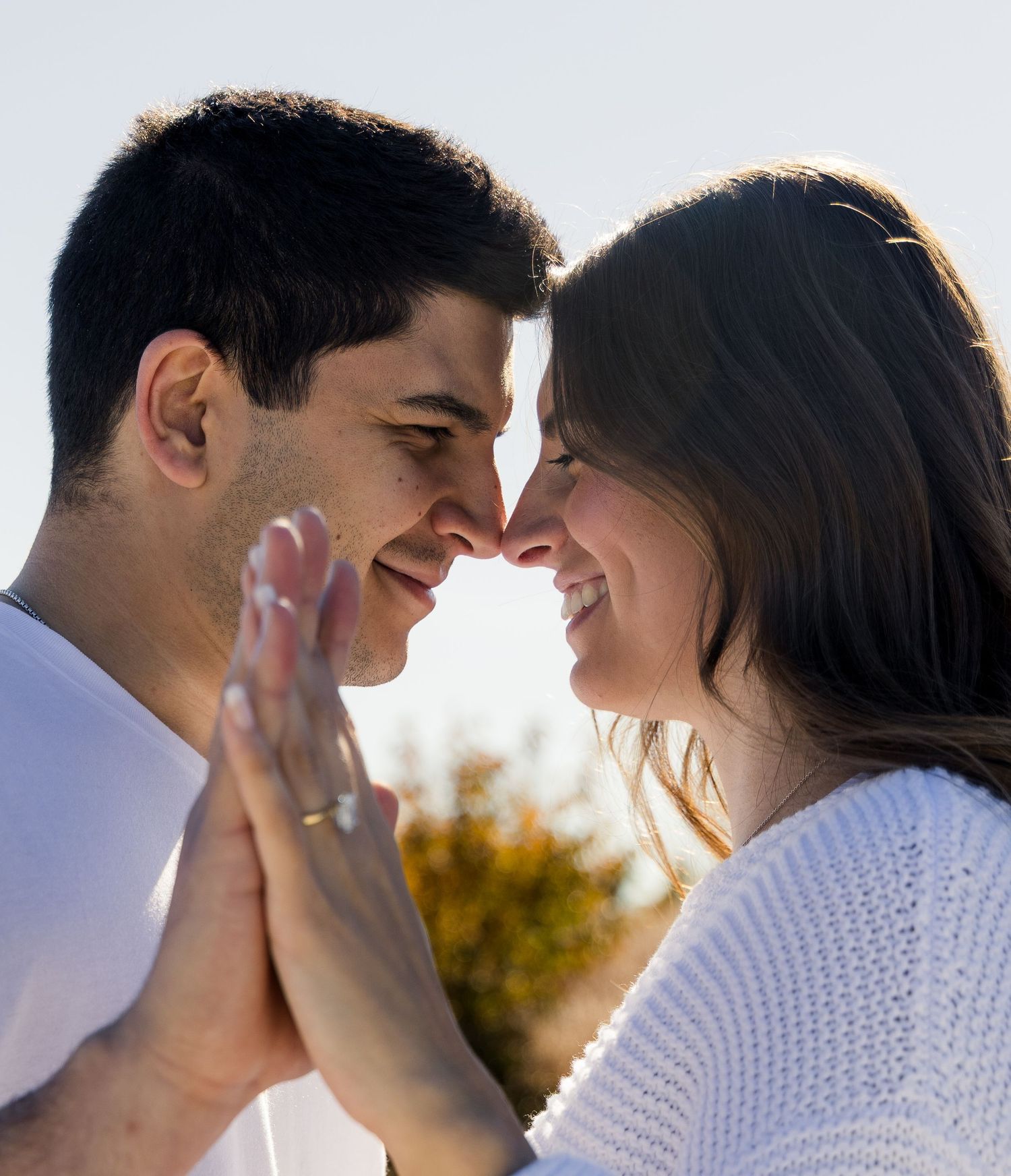 Warm, intimate outdoor couples photoshoot with smiling couple, touching foreheads and showing engagement ring