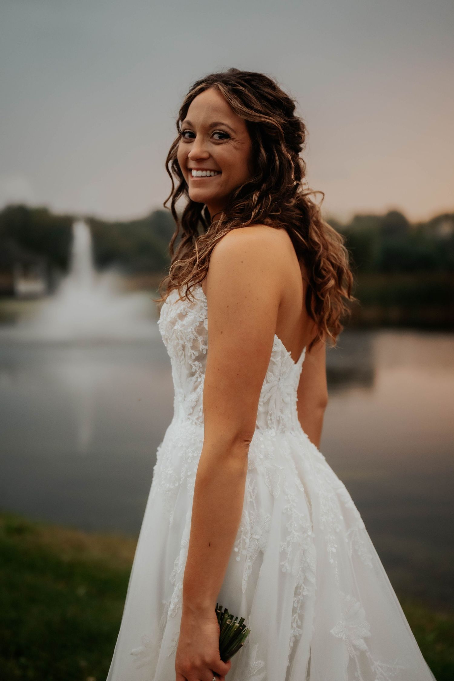 Radiant bride in lace ball gown posing by fountain at sunset.