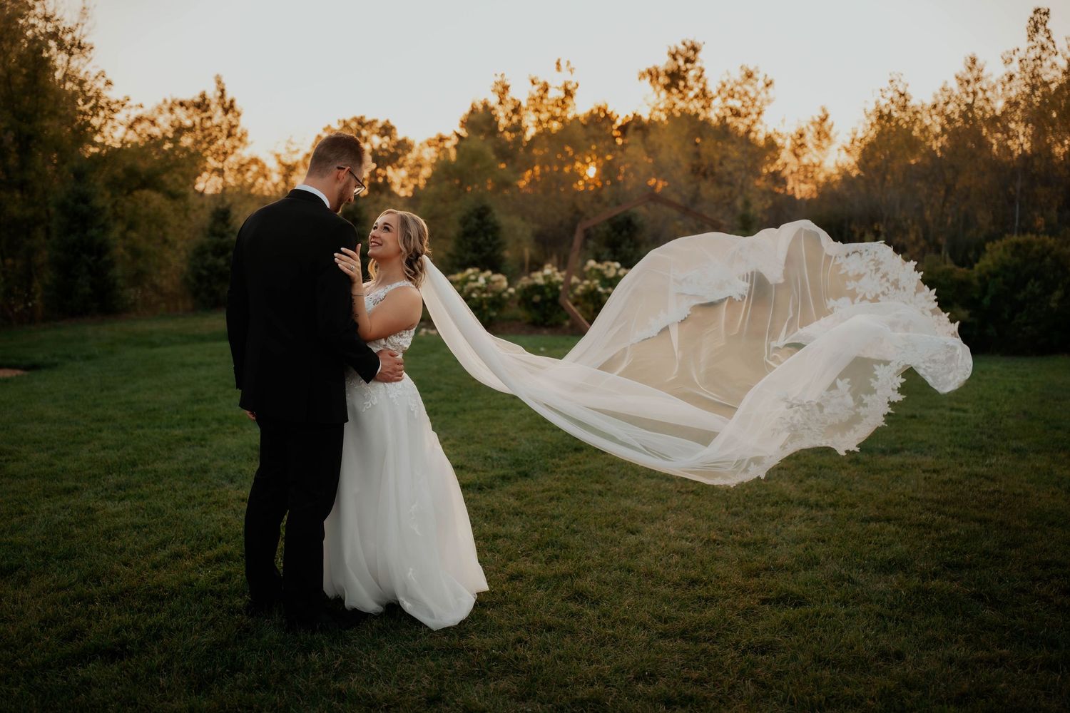Bride and groom standing close as flowing wedding veil billows in the wind during golden hour.