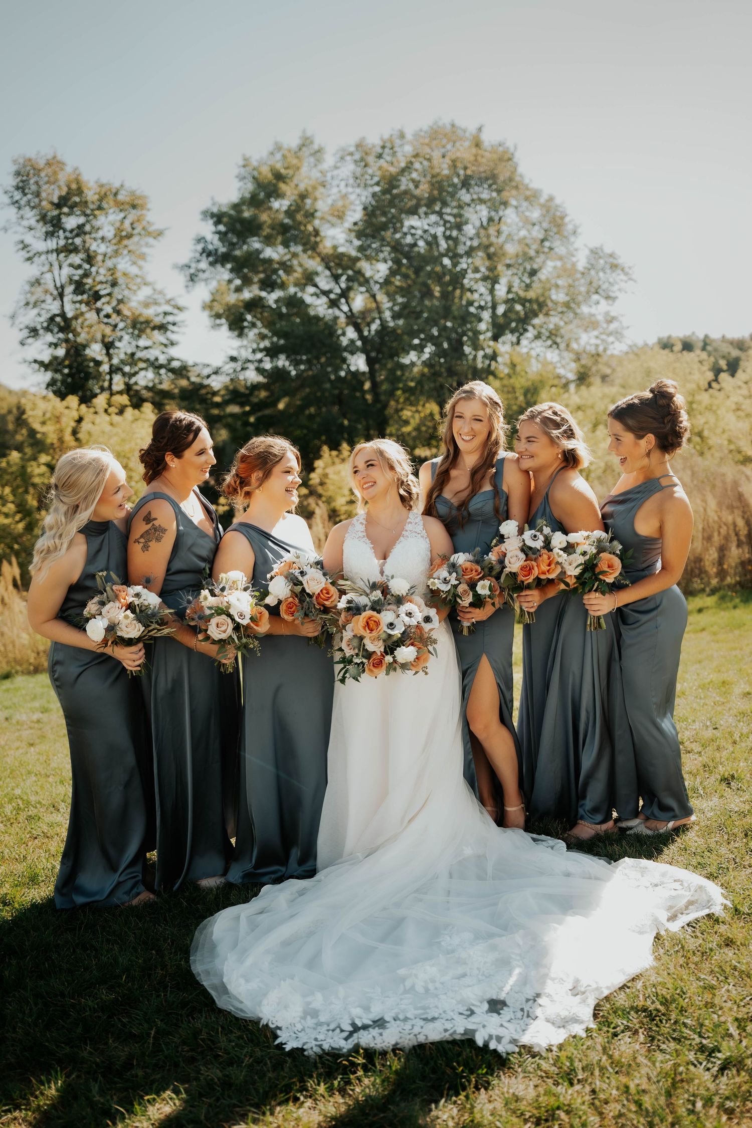 Bride surrounded by six bridesmaids in slate blue gowns holding orange and white bouquets outdoors.