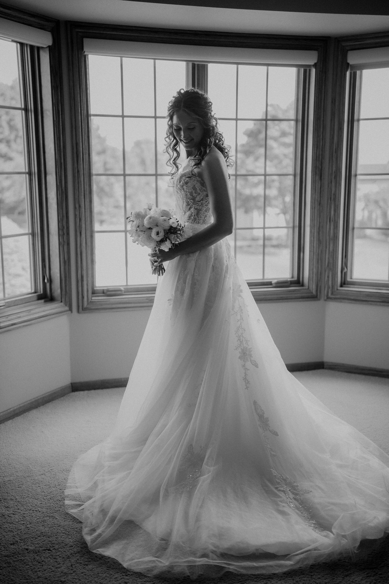Bride in elegant white lace ball gown holding bouquet standing before bay windows in black and white.