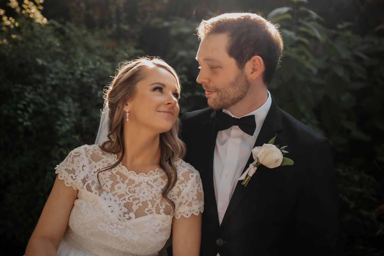 Romantic bride and groom portrait in golden sunlight among lush greenery.