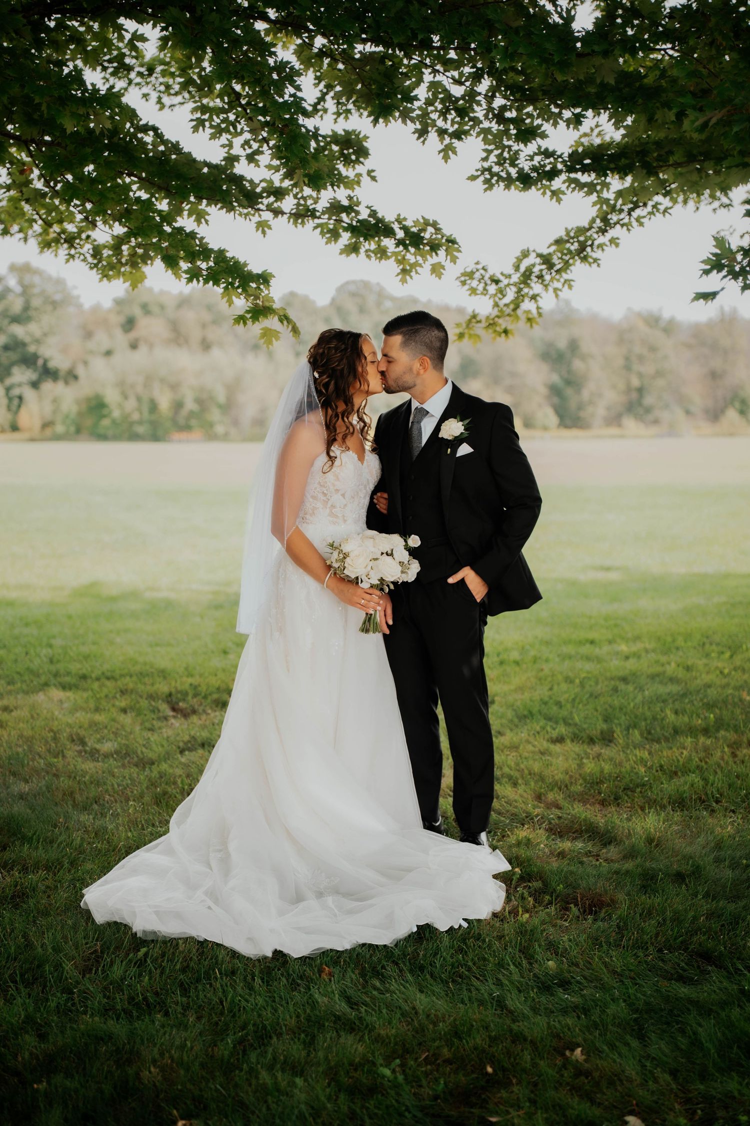Bride and groom kissing under a leafy tree in a lush green field during outdoor wedding portraits.