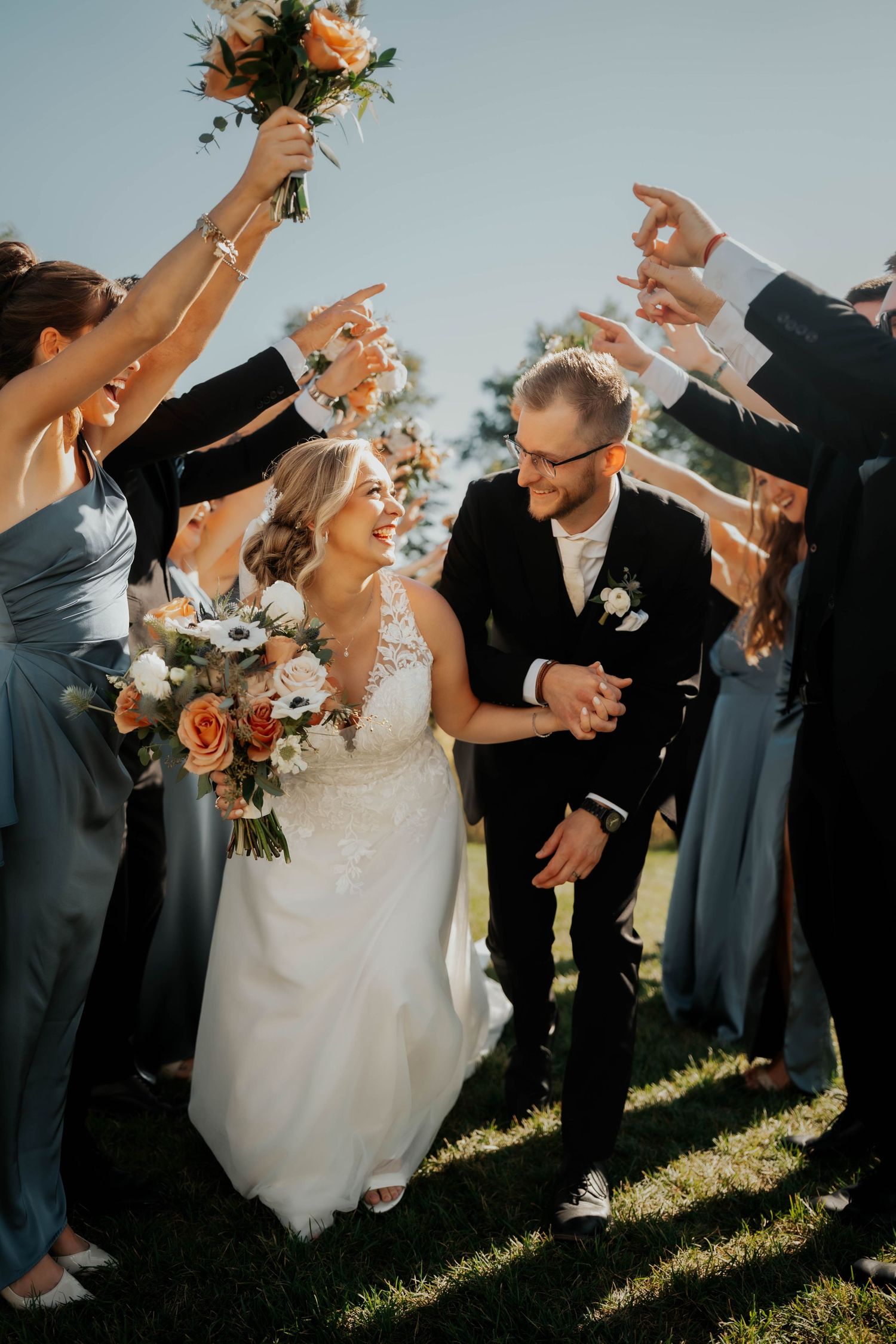 Bride and groom walk through tunnel of raised hands and bouquets held by cheering wedding party outdoors.