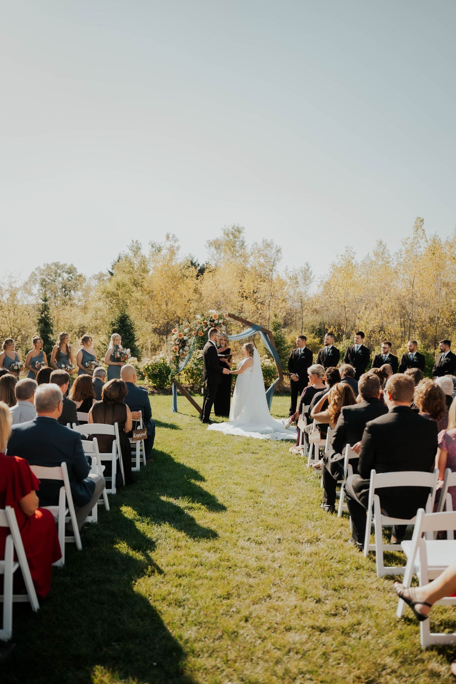 Outdoor wedding ceremony with guests seated in white chairs as couple exchanges vows under floral arch.