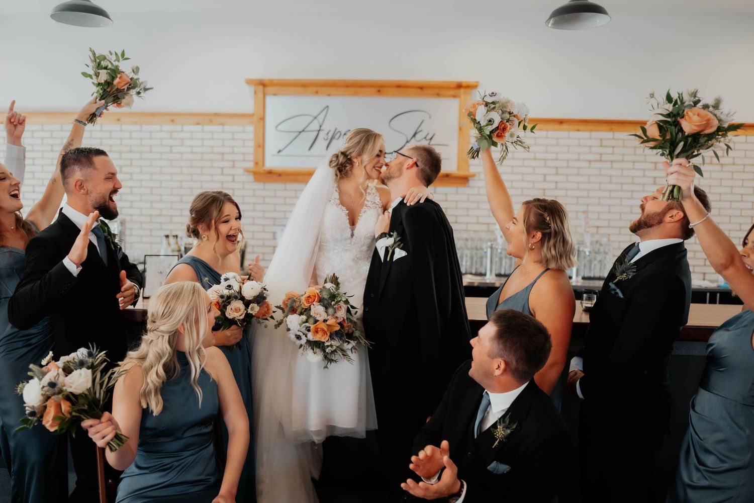 Wedding party celebrates as bride and groom kiss indoors, raising bouquets and cheering joyfully.