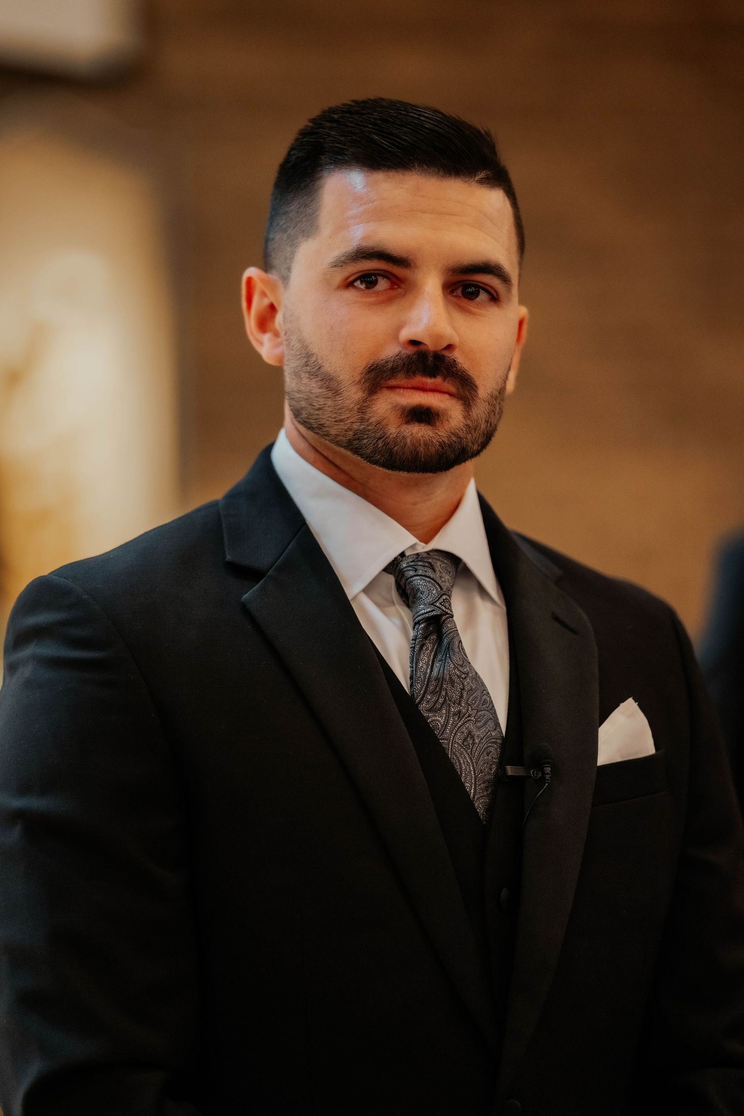 Handsome groom in black suit with paisley tie and white pocket square posing at wedding ceremony.