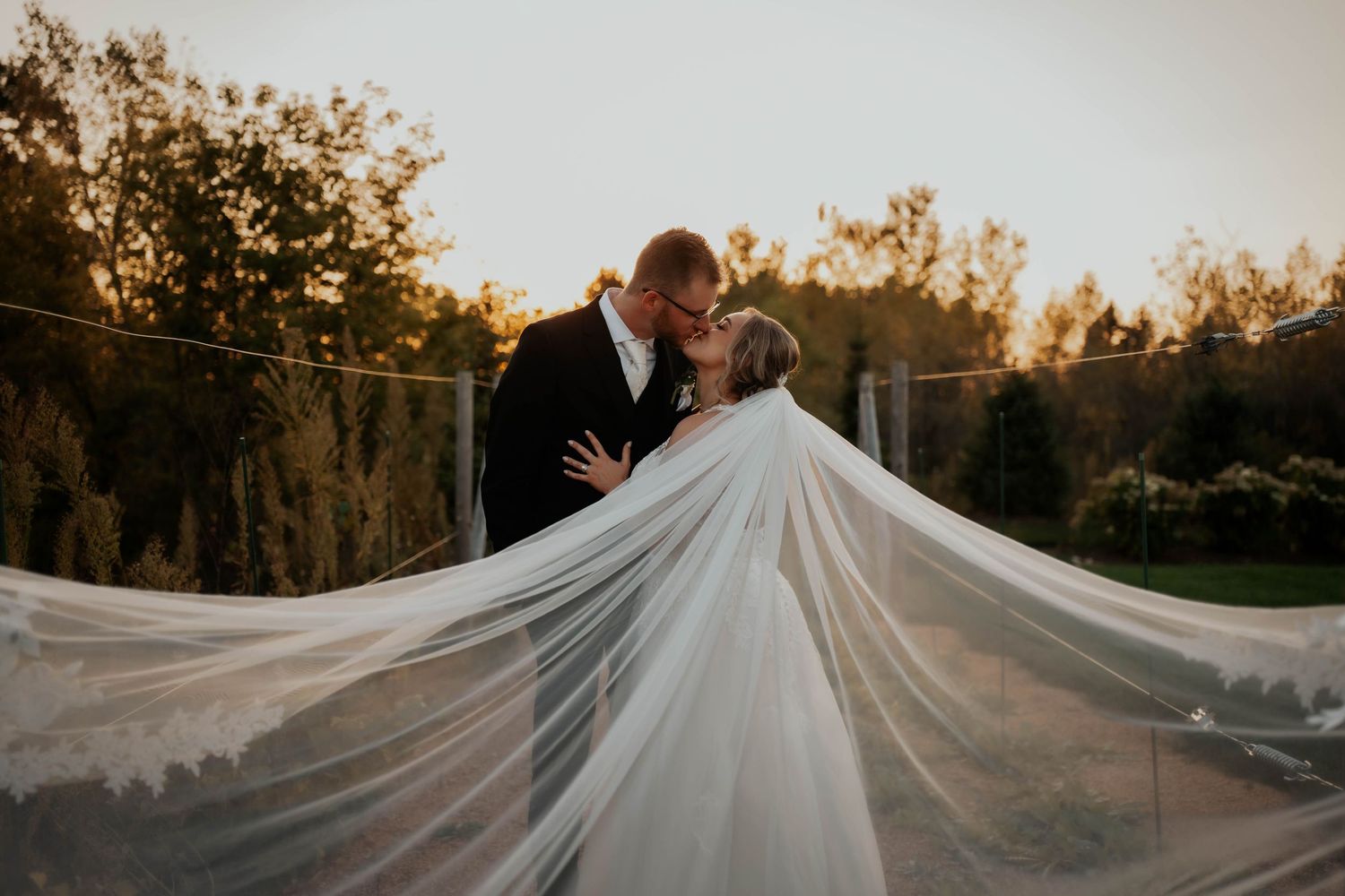 Bride and groom sharing a romantic kiss at golden hour, wedding veil dramatically spread wide in the breeze.