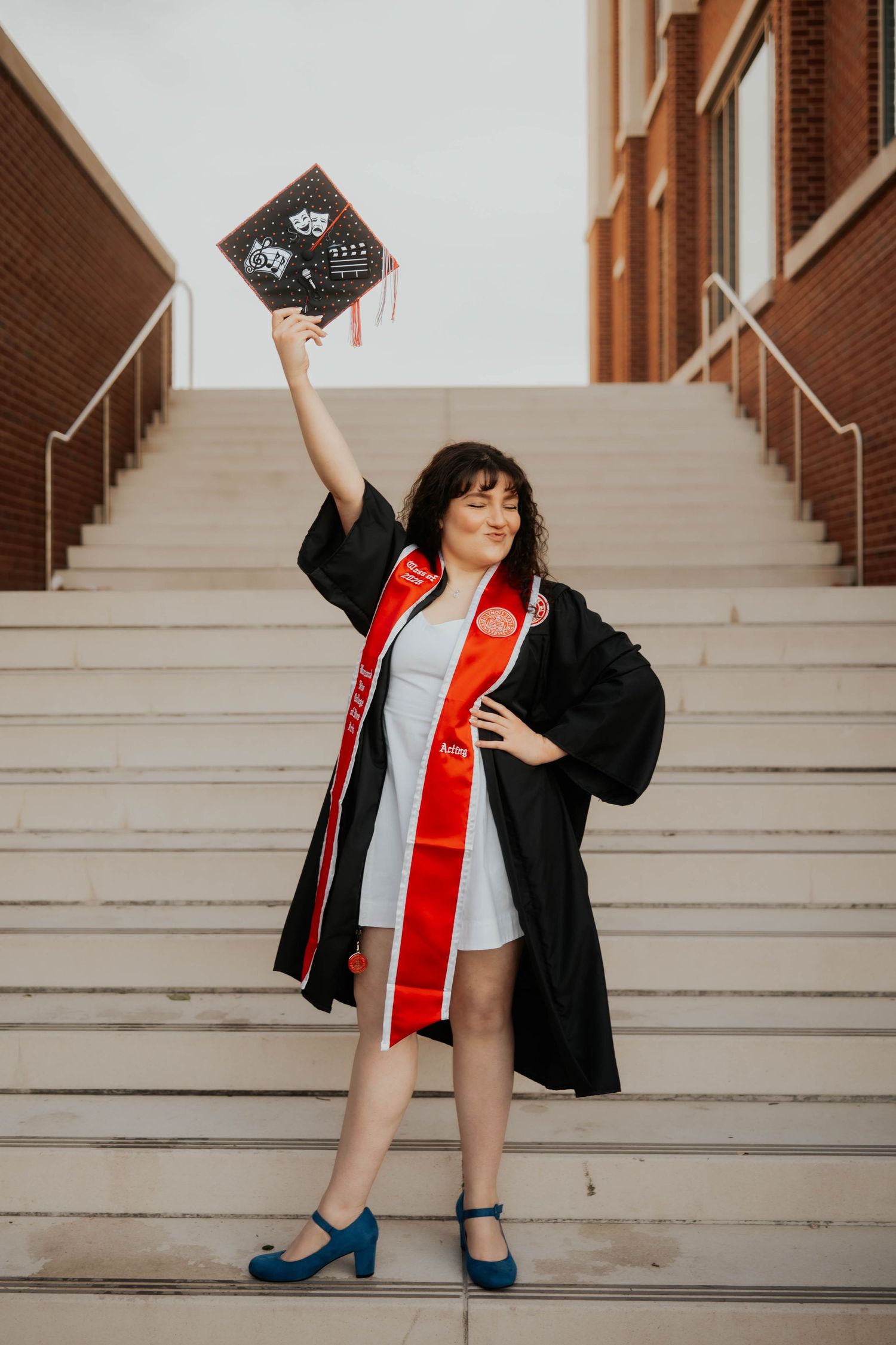 Graduate in black cap and gown with red sash celebrates on university steps, raising decorated cap high.