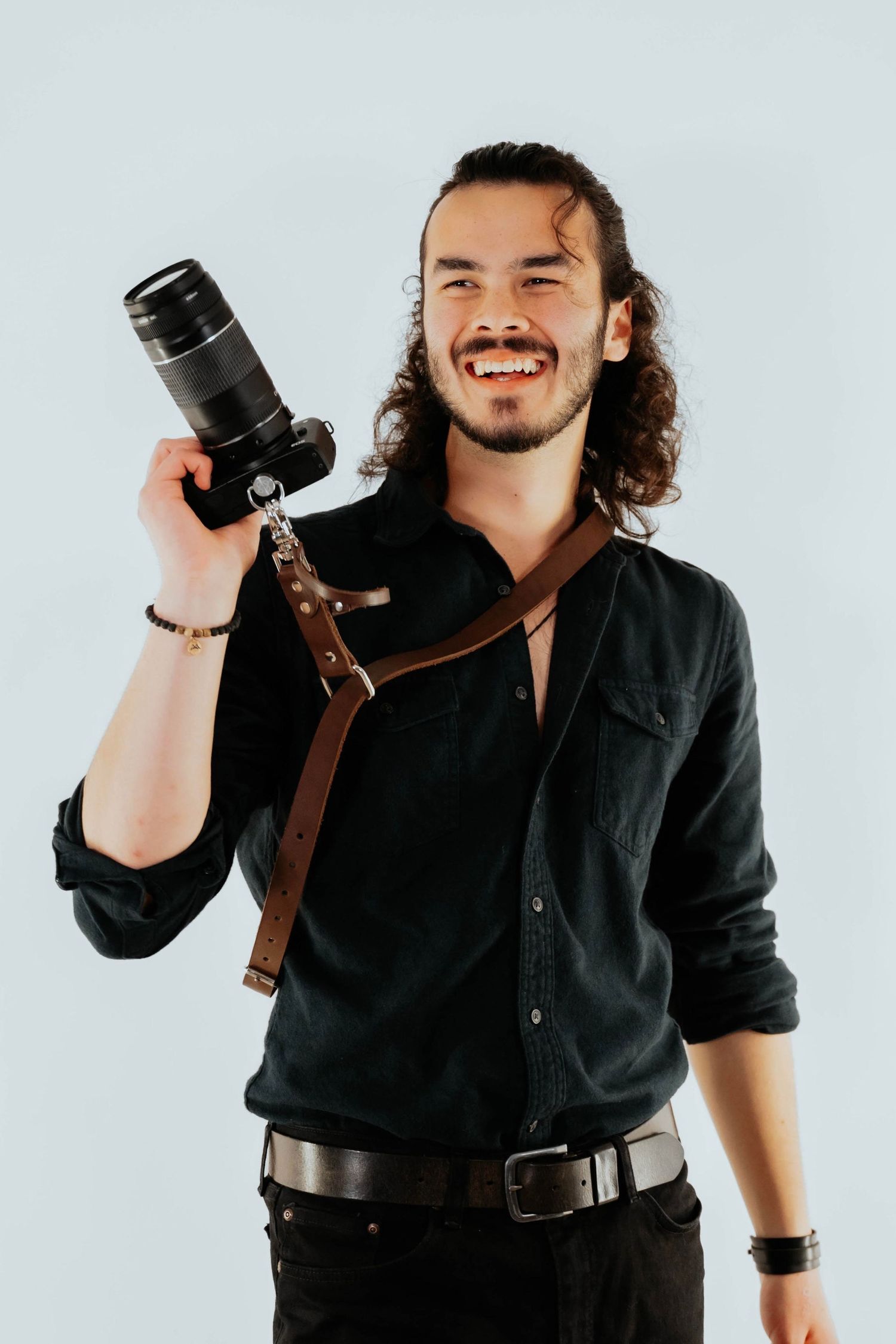 Happy male photographer with long curly hair holding a professional camera with leather strap in a studio portrait.