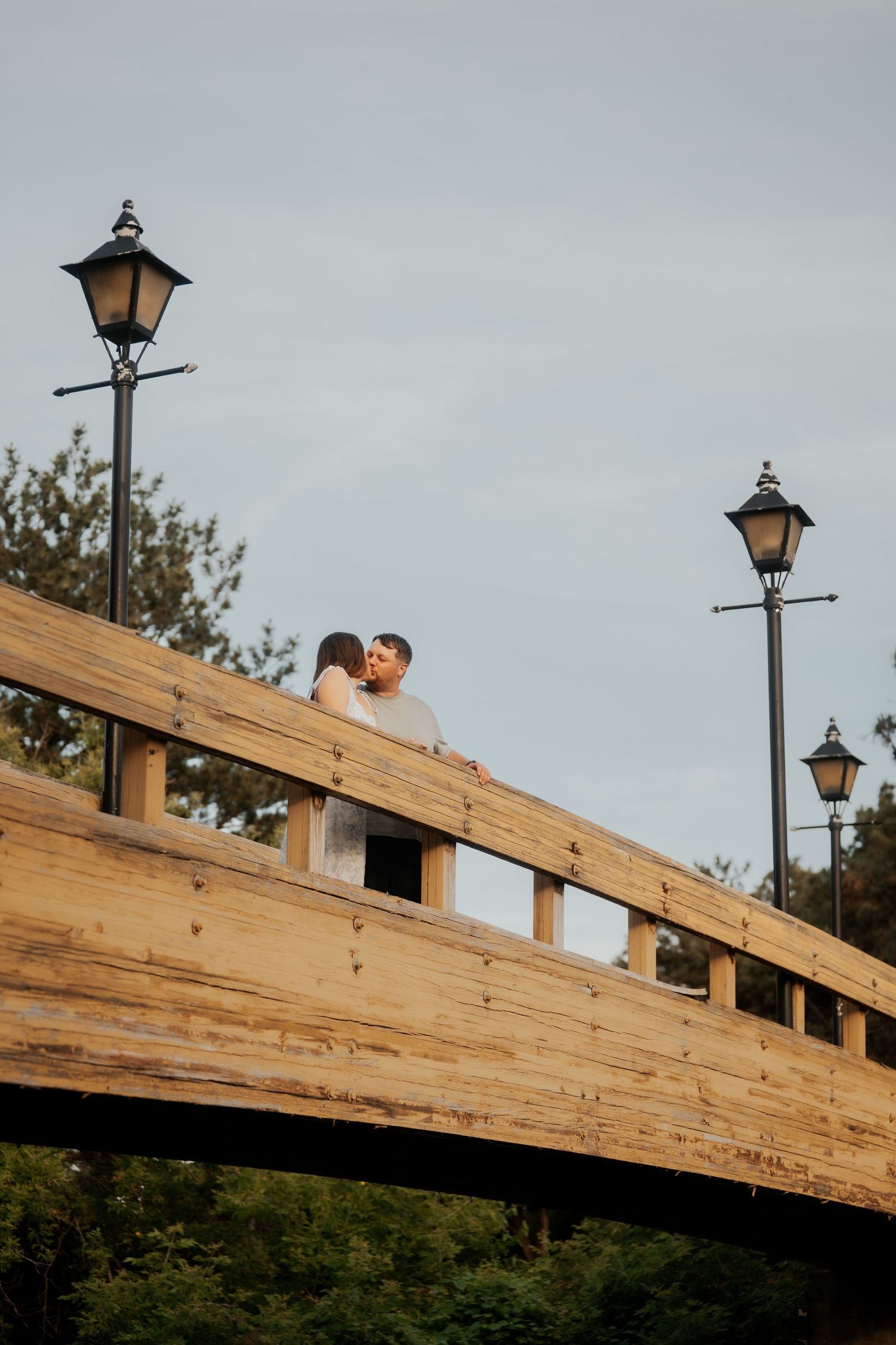 Couple sharing a romantic kiss on a wooden bridge flanked by vintage lamp posts.