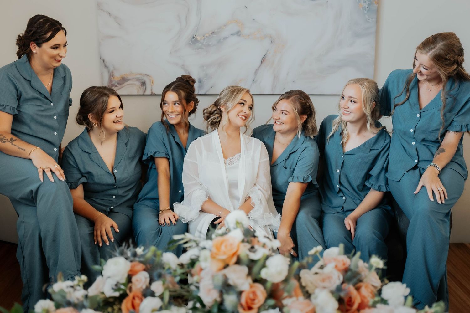 Bride in white robe laughs joyfully with bridesmaids wearing matching teal jumpsuits before the wedding.