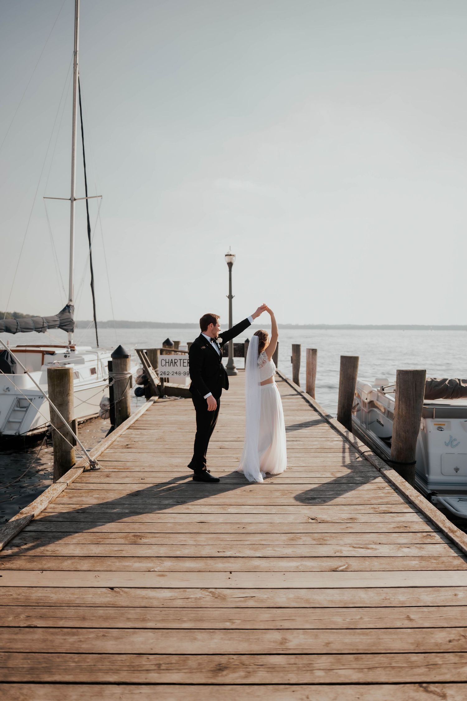 Couple dancing joyfully on wooden pier with sailboats and open water.