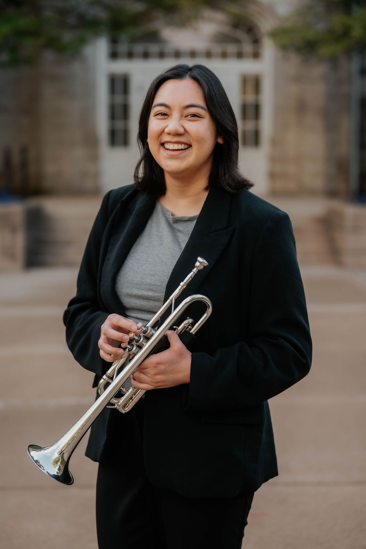 Smiling young woman in black blazer holds a silver trumpet outdoors in a courtyard setting.