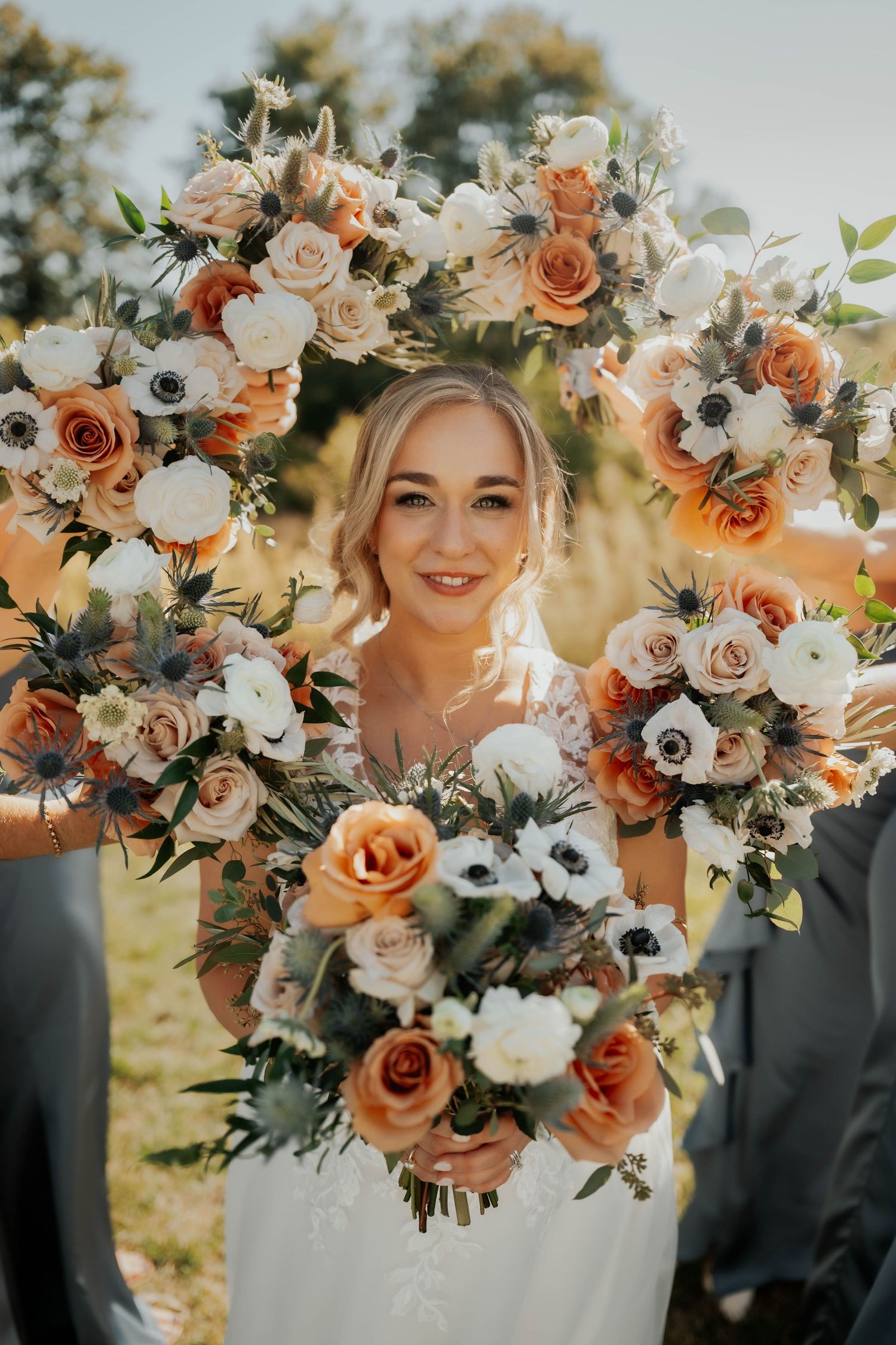 Bride framed by bridesmaids holding orange, white, and peach floral bouquets with anemones and roses.
