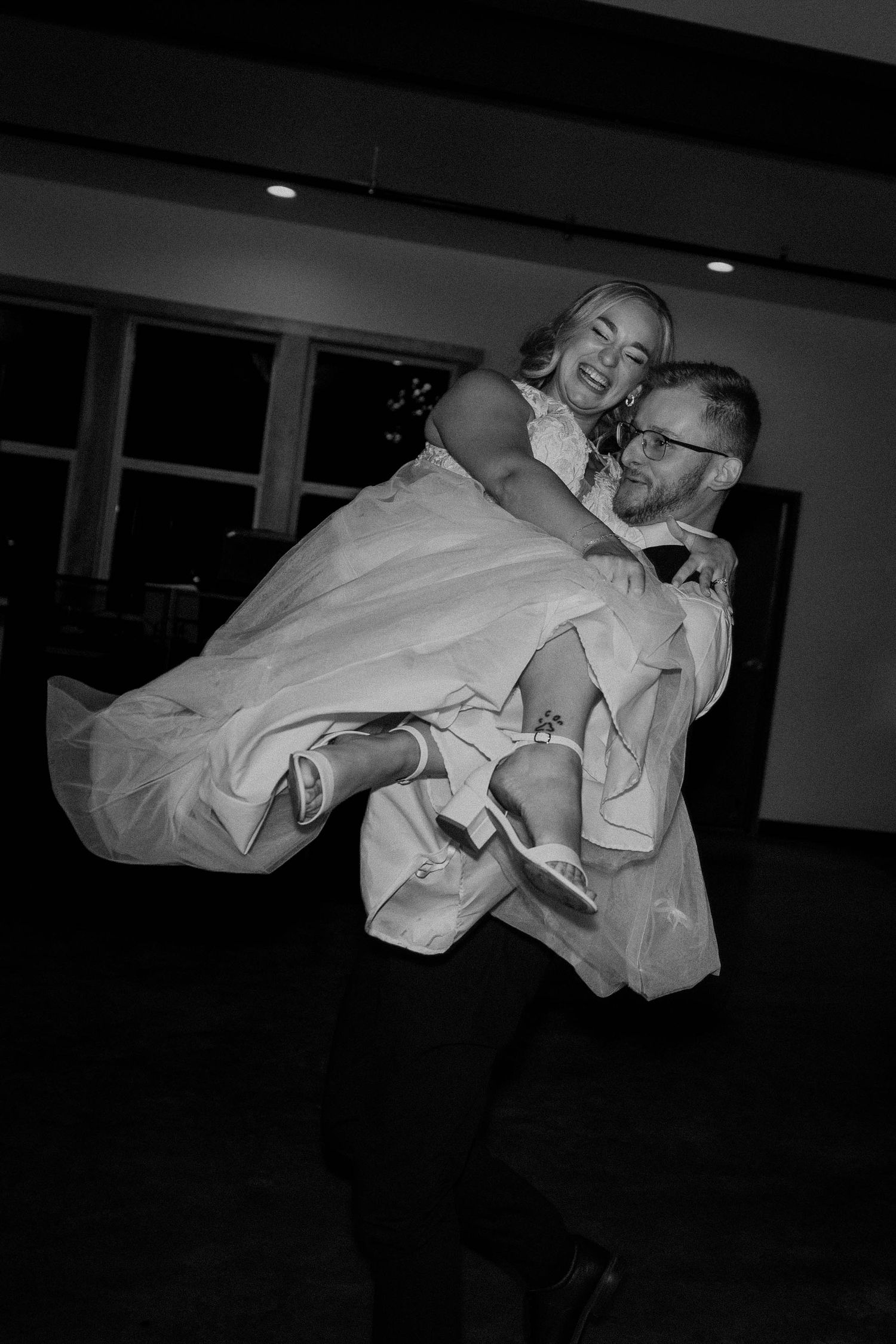 Groom joyfully carries bride on his back during wedding reception dance in black and white photo.
