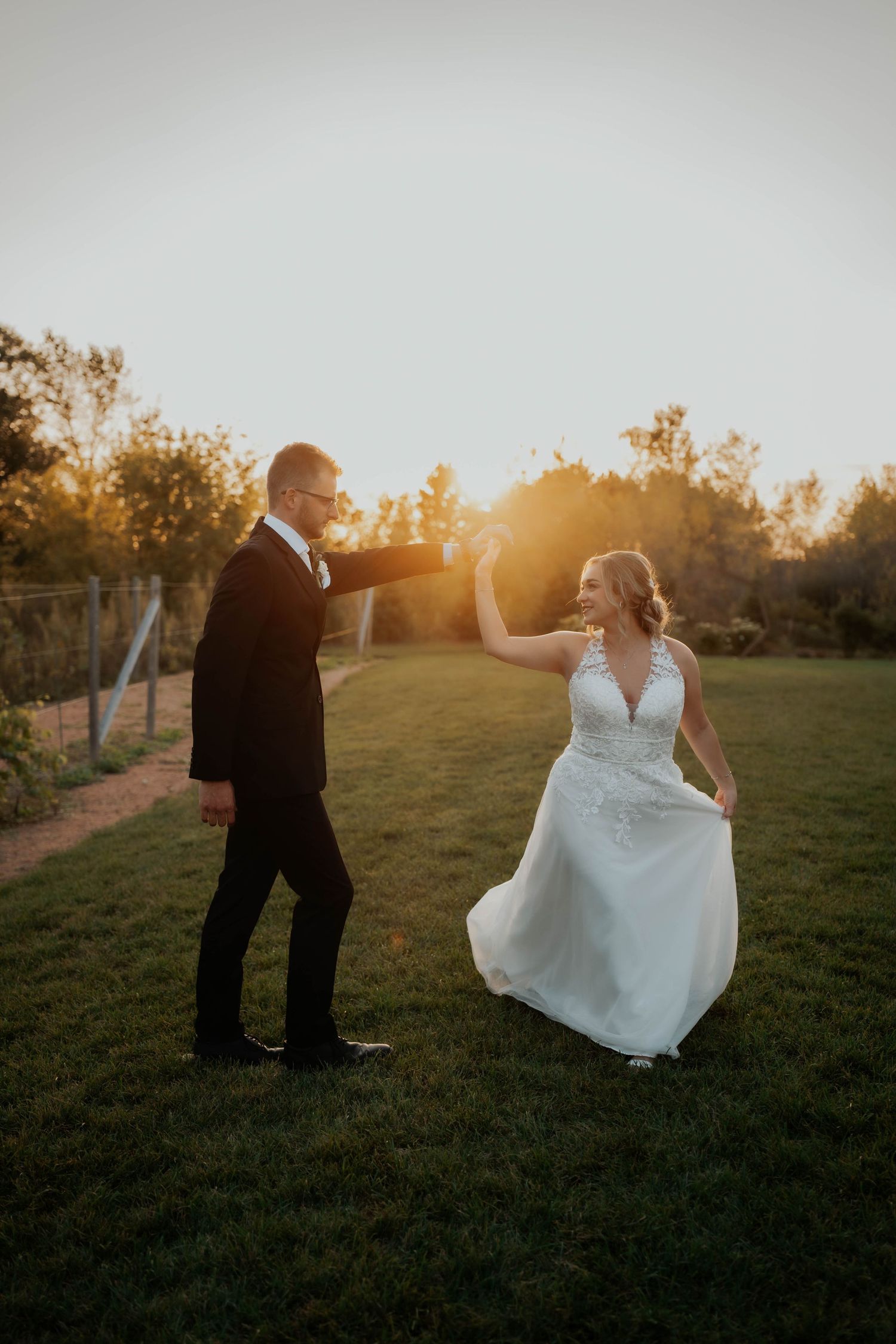 Groom twirling bride in white lace gown on green lawn during stunning sunset at outdoor wedding.