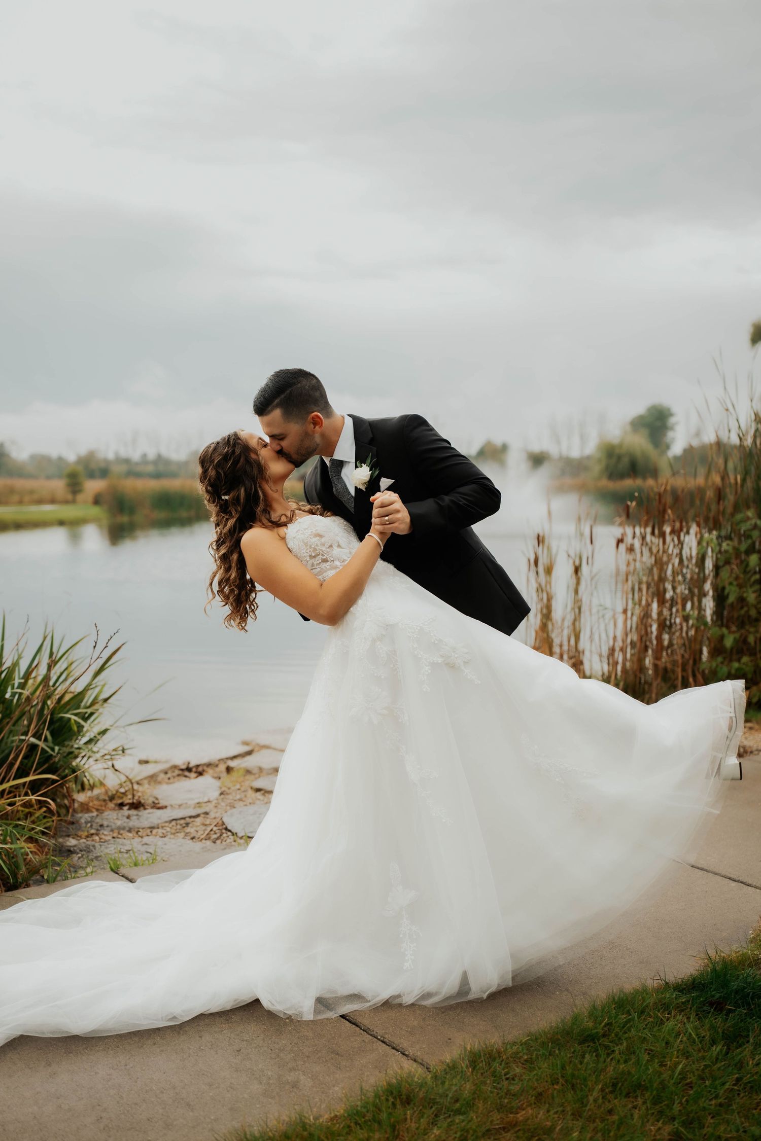 Groom dipping bride in flowing white gown for a kiss beside a scenic pond on wedding day.