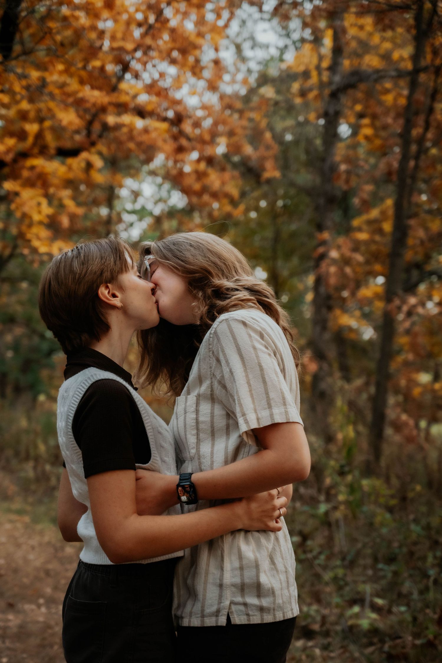 Two people share a romantic kiss surrounded by colorful autumn foliage in a forest.