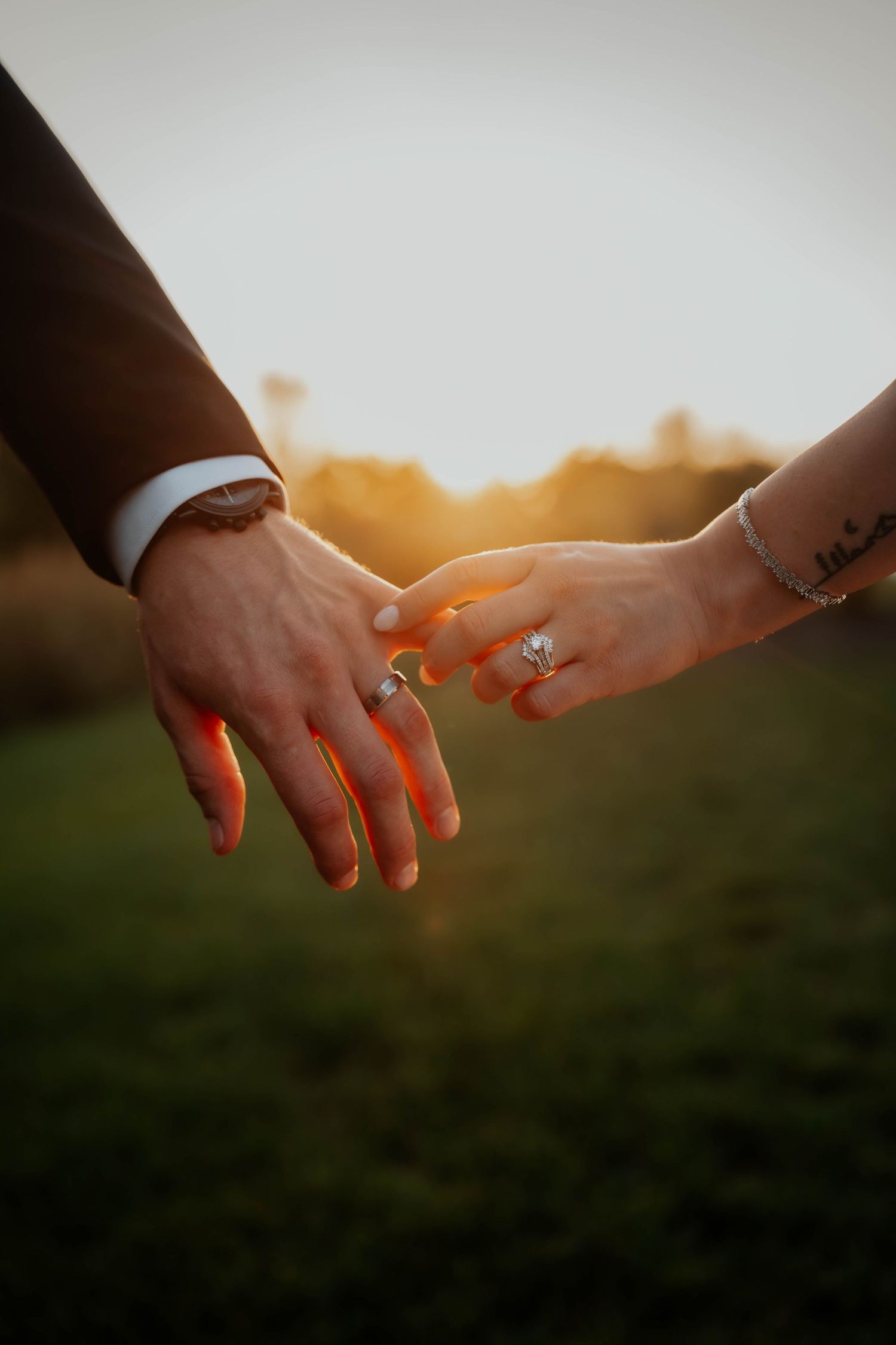 Close-up of bride and groom holding hands at sunset, showcasing wedding rings and bride's wrist tattoo.