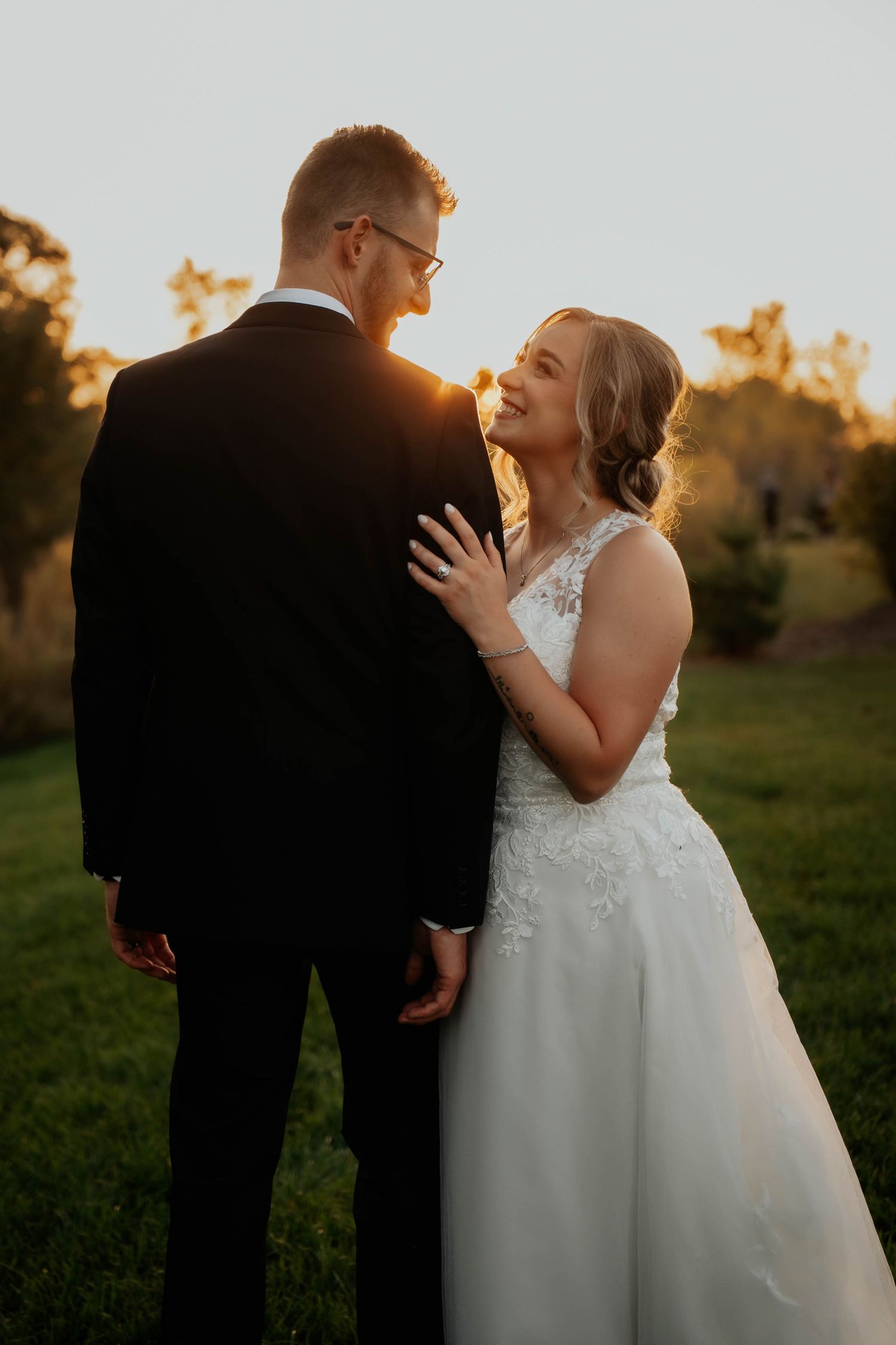 Bride in lace wedding gown smiling at groom during golden hour sunset in outdoor garden setting.