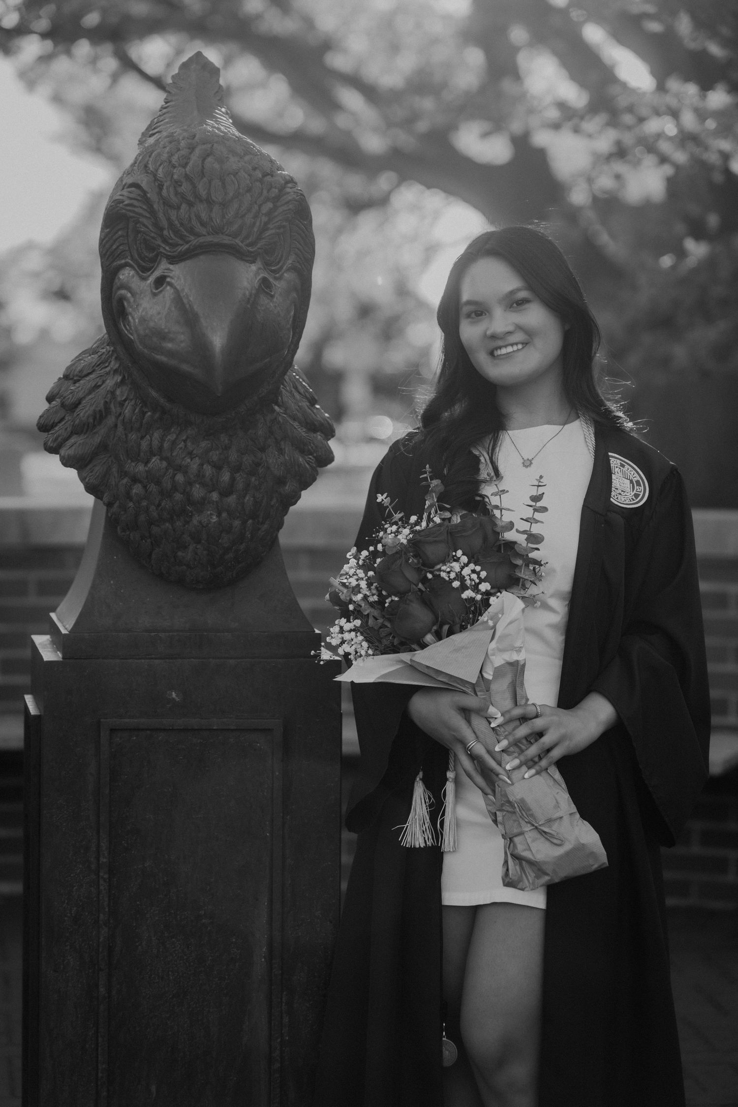 Black and white photo of smiling graduate in cap and gown holding flowers beside eagle statue.