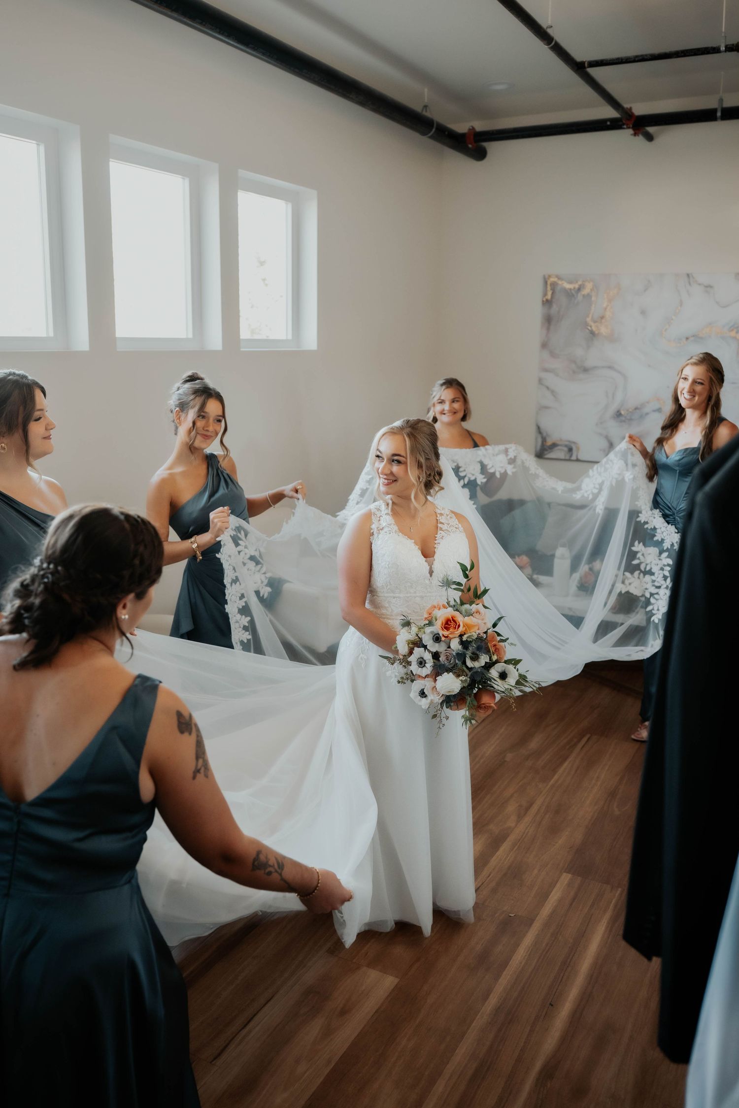 Bridesmaids in slate blue gowns hold out bride's lace-trimmed veil as she holds bouquet indoors.