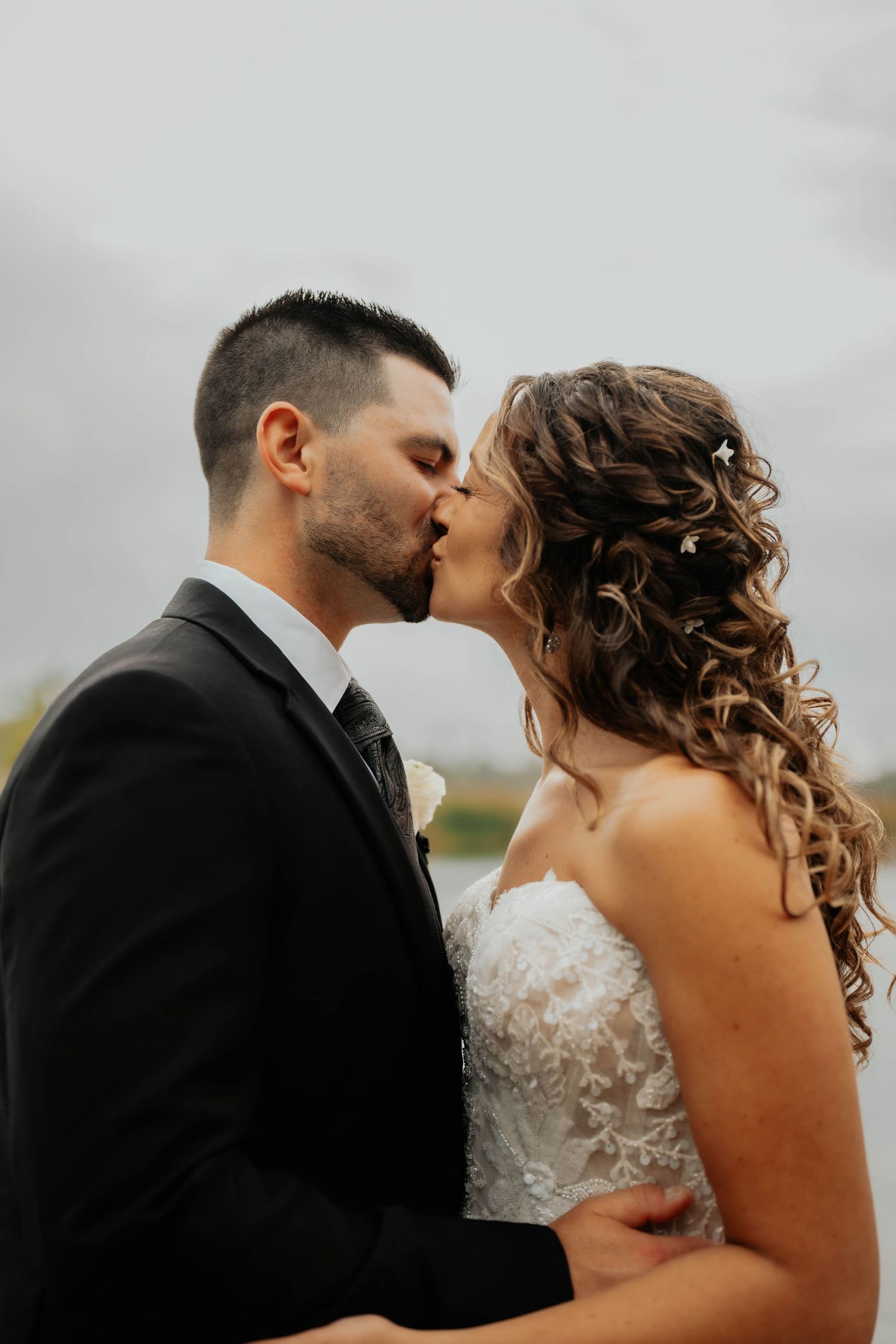 Bride and groom sharing a romantic kiss outdoors with overcast sky and water in the background.