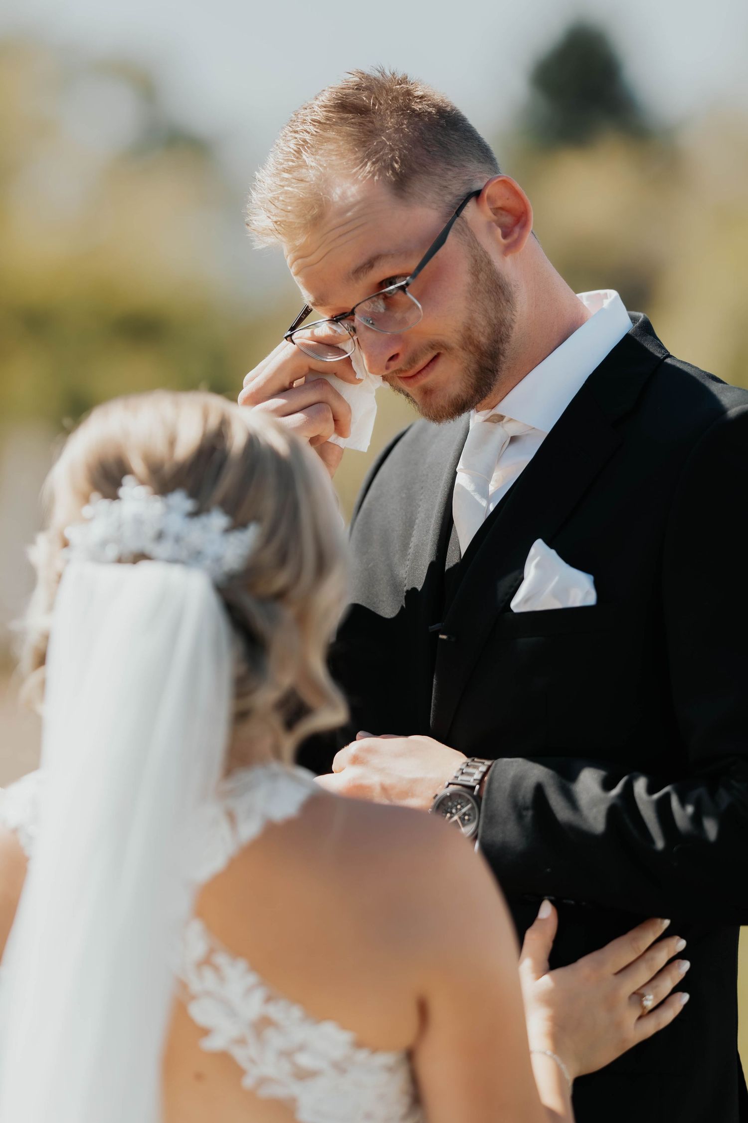 Emotional groom in black suit wiping tears while seeing his bride during outdoor wedding ceremony.
