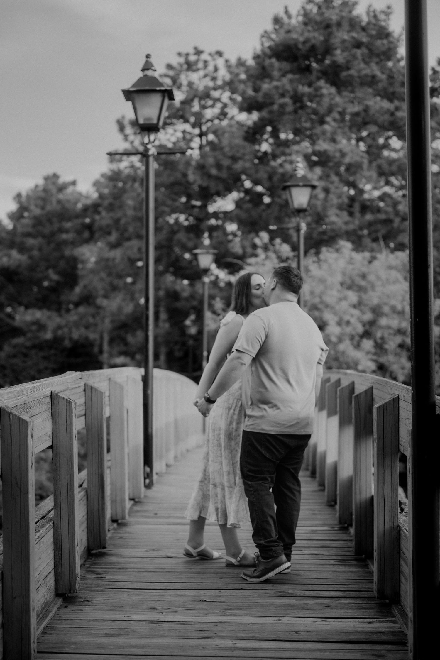 Black and white rear view of couple strolling on wooden bridge with lanterns and trees behind them.