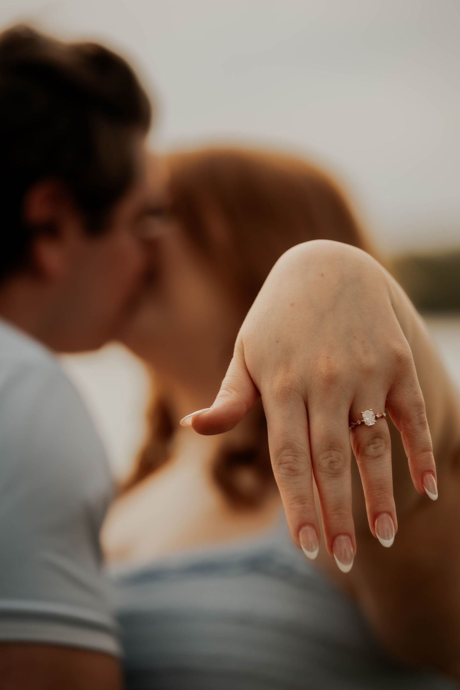 Close-up of engagement ring on woman's hand as couple kisses in background.