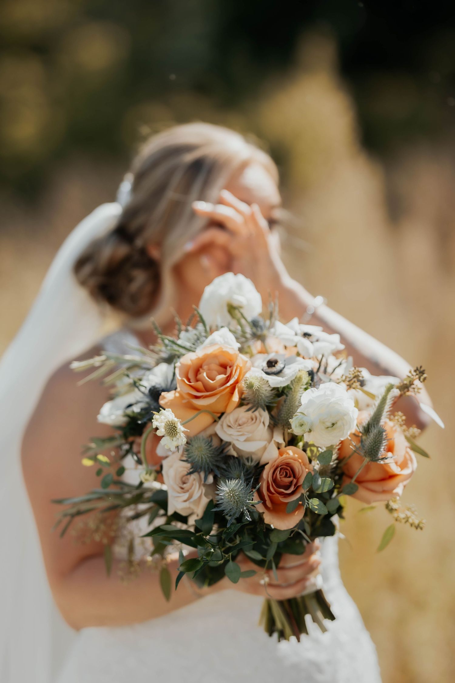 Bride holding a stunning bouquet of orange roses, white anemones, and thistles in a sunny field.