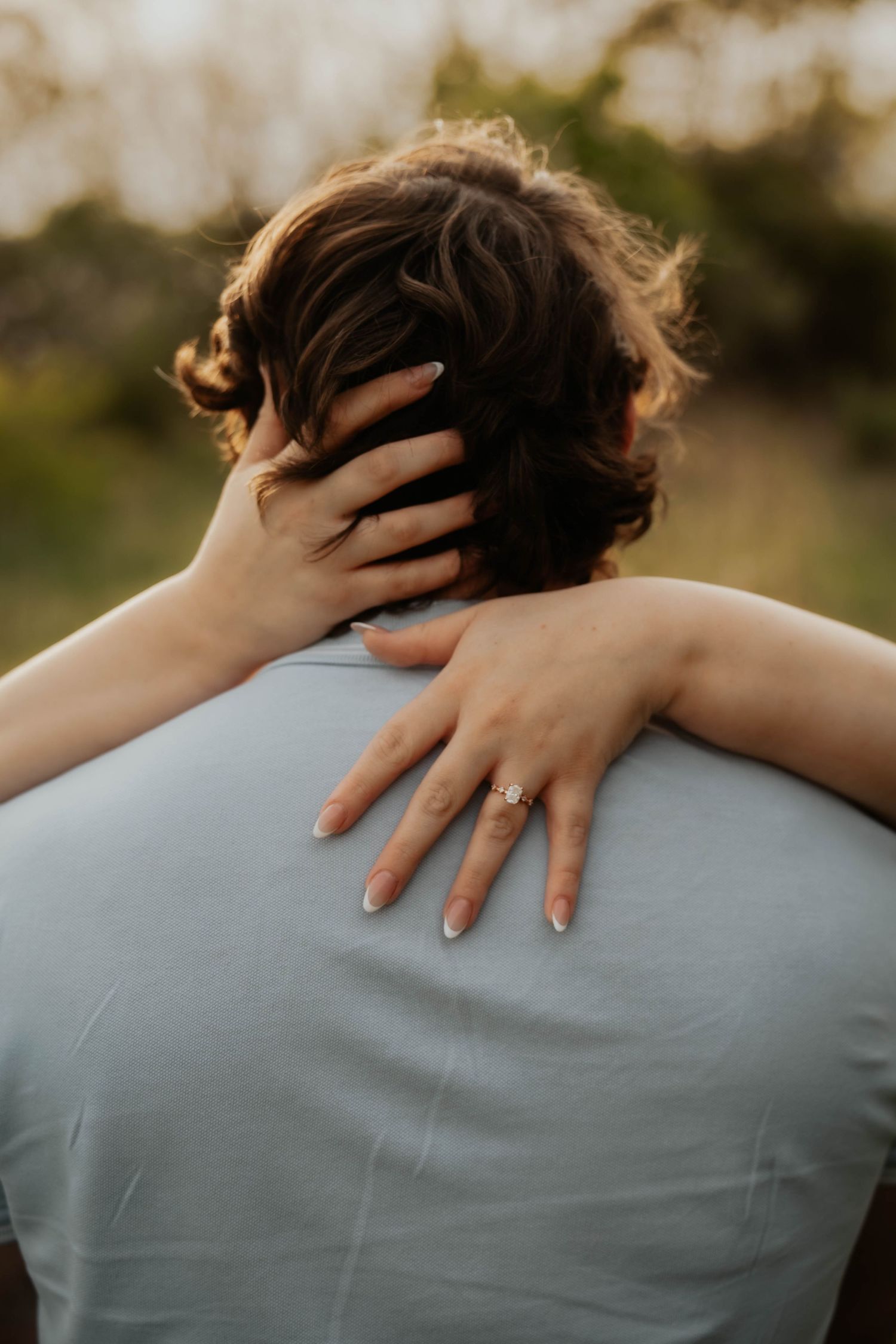 Woman with engagement ring embracing man from behind outdoors.