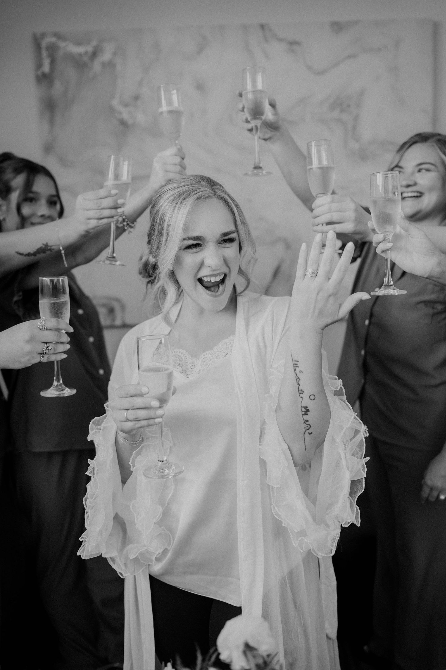 Excited bride in white robe toasts champagne with bridesmaids in black and white celebration photo.