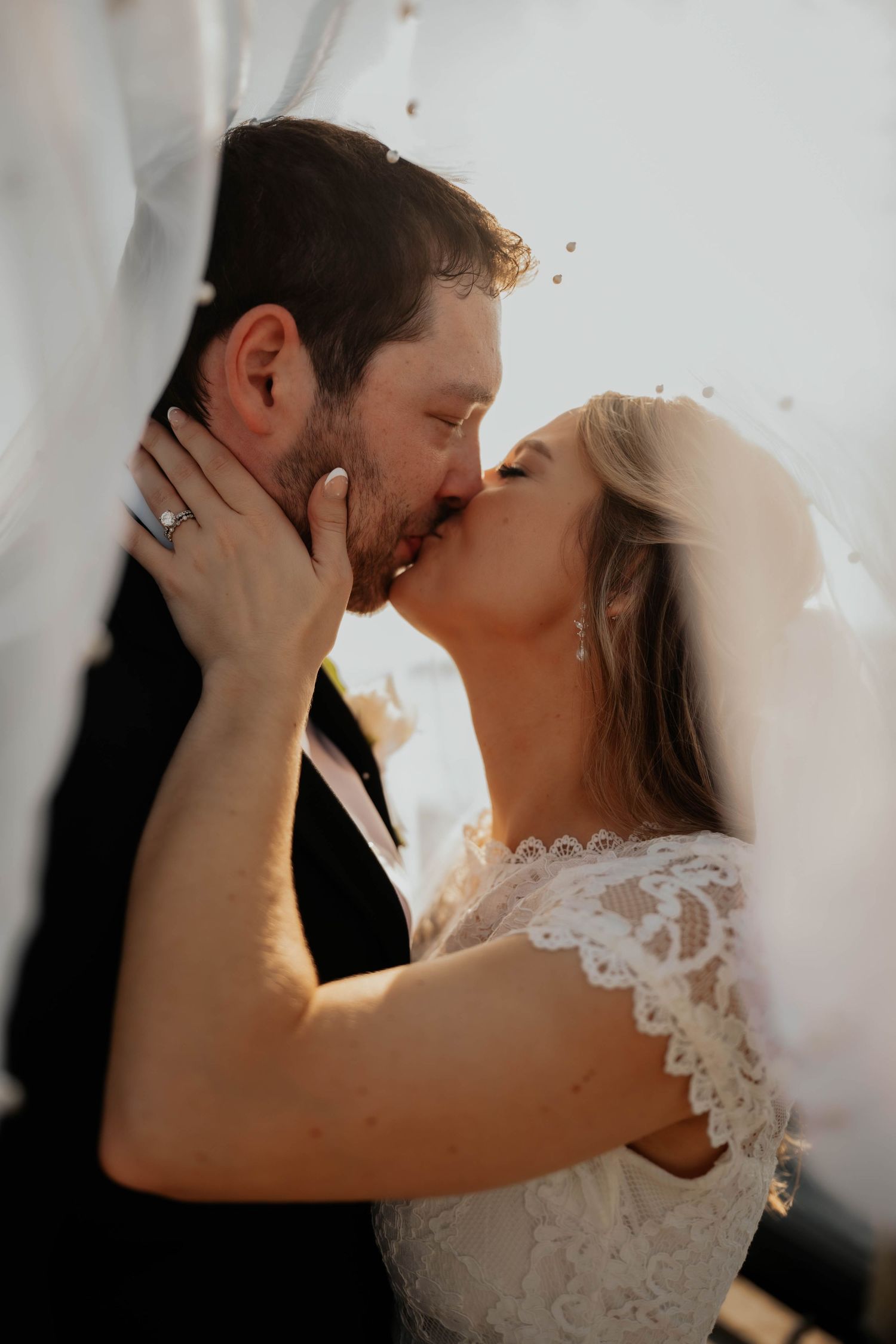 Newlyweds sharing passionate first kiss under flowing white veil in sunlight.