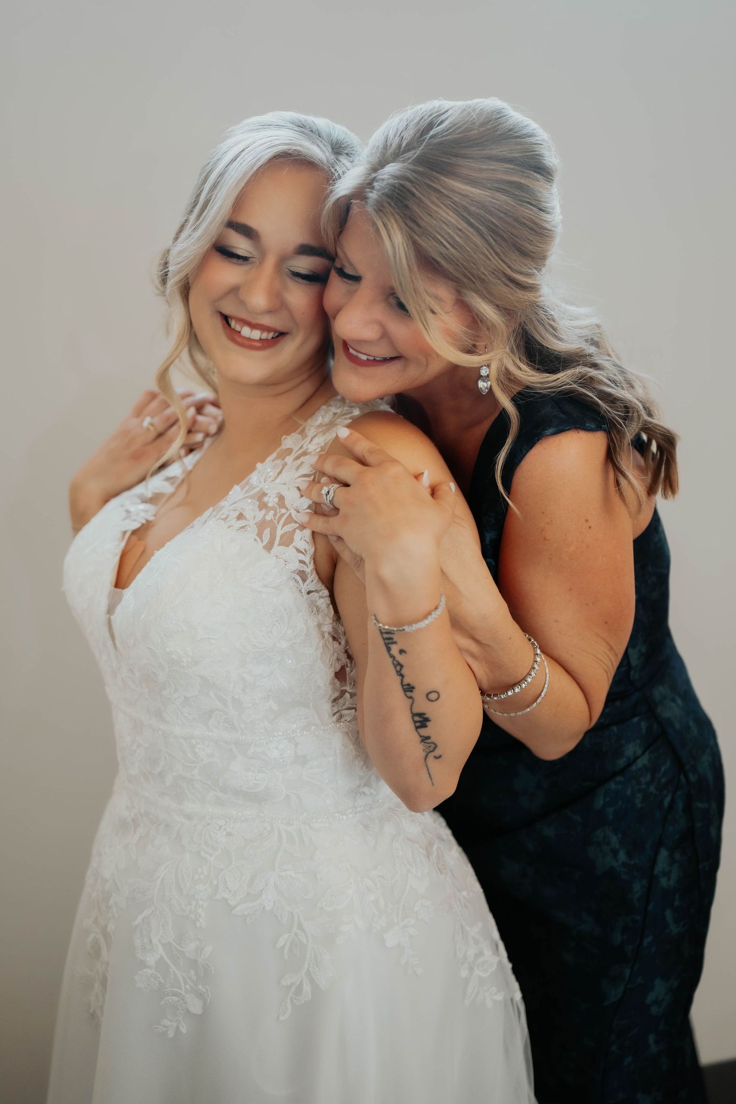 Radiant bride in lace gown shares tender embrace with smiling woman in elegant dark dress.