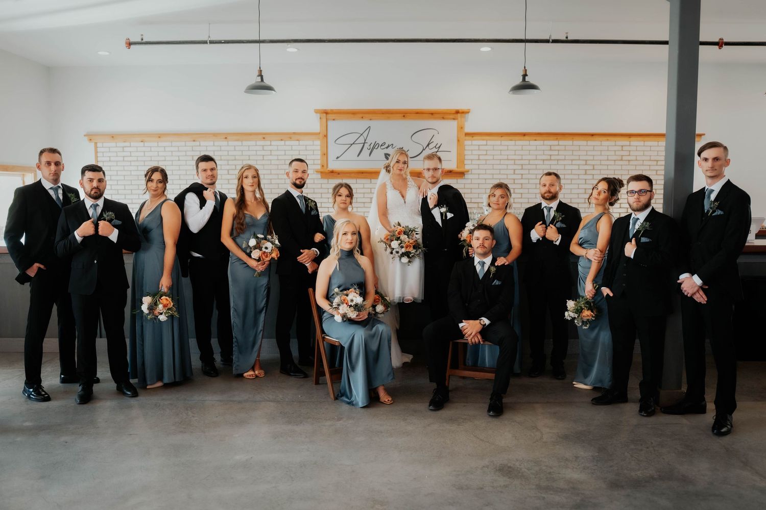 Large wedding party poses indoors at Aspen venue in slate blue bridesmaid gowns and black tuxedos.