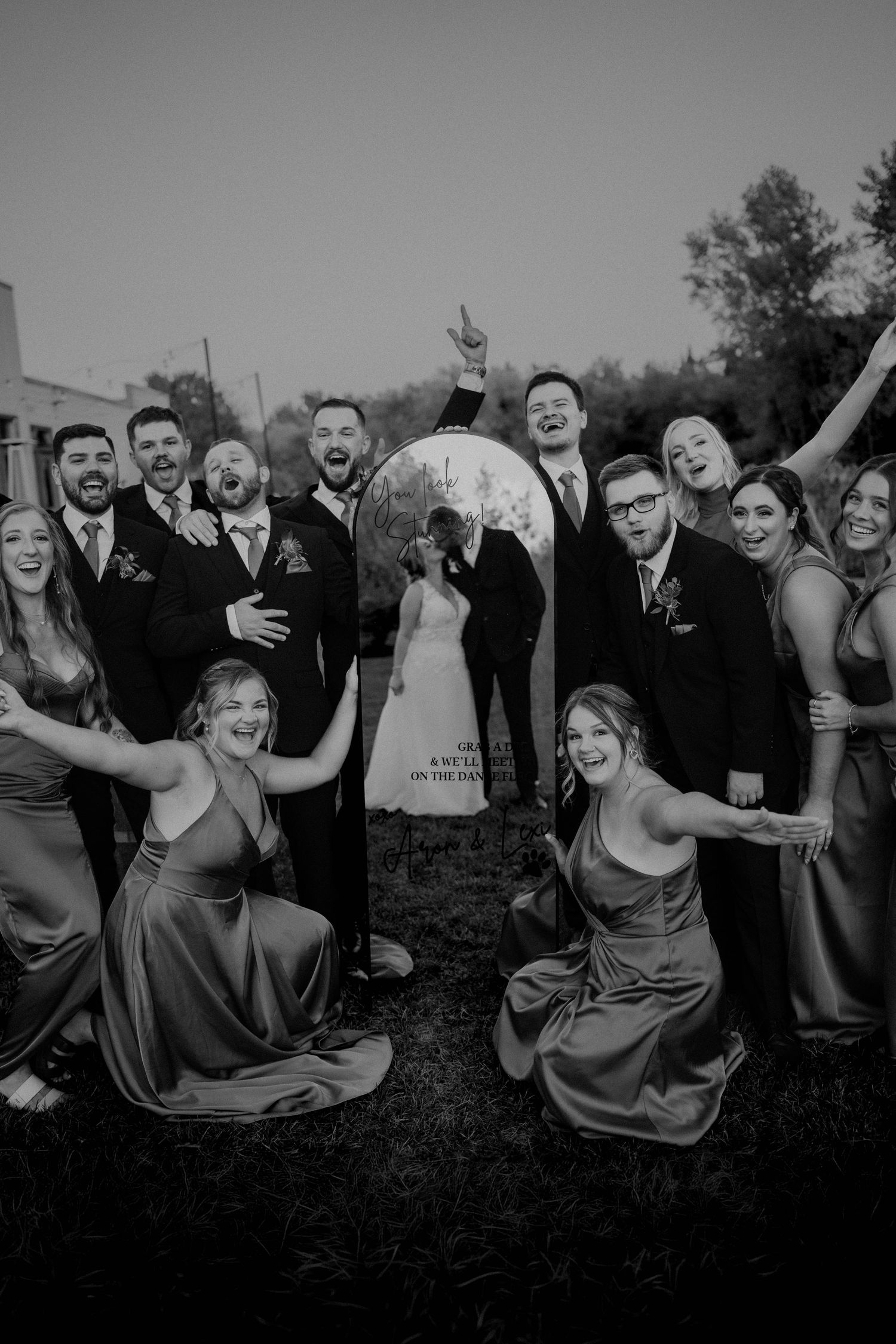 Black and white wedding party photo with bride and groom kissing near globe sculpture surrounded by cheering bridesmaids and groomsmen.