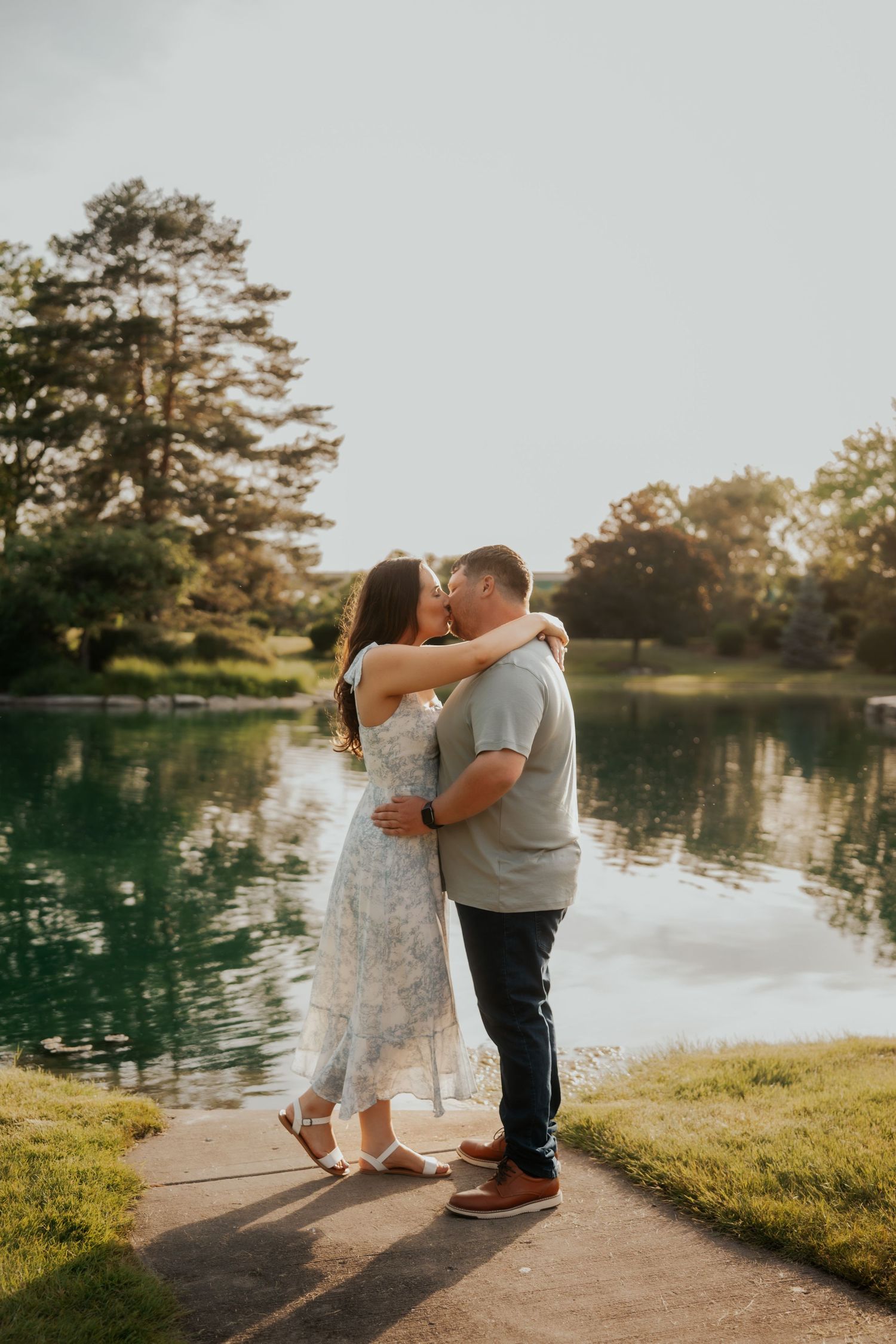 Engaged couple sharing a romantic kiss beside a serene reflective pond at sunset.