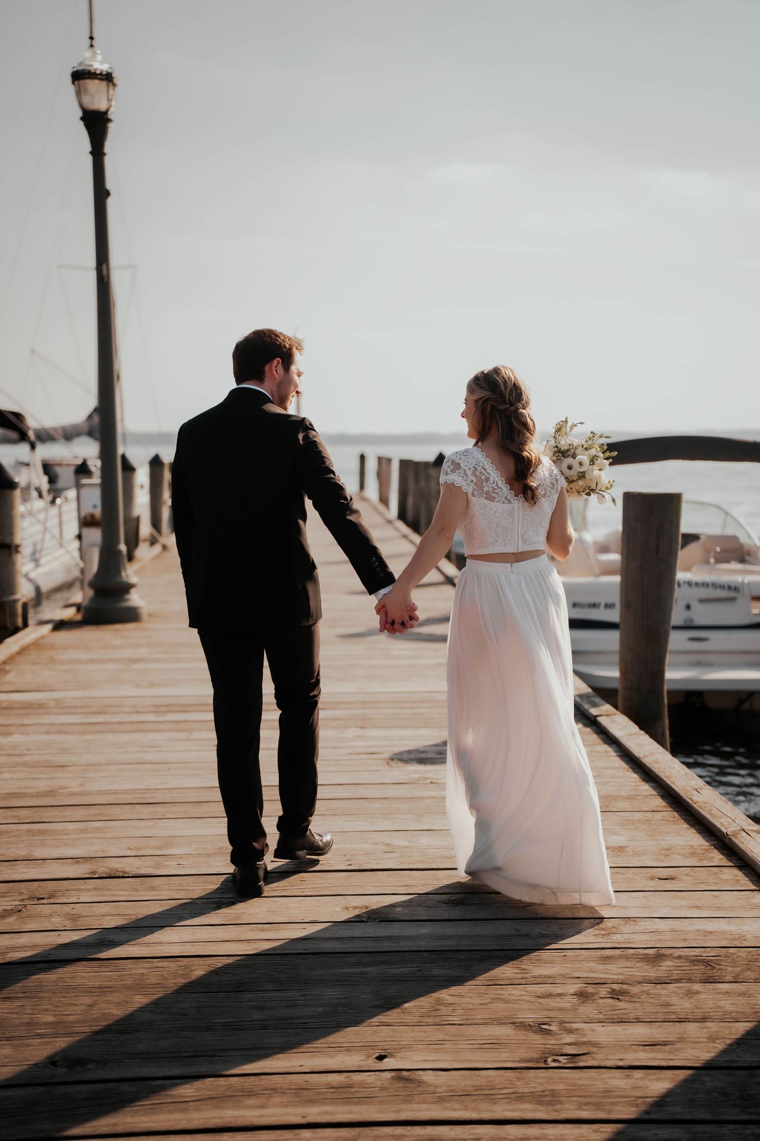 Bride and groom walking hand in hand along sunny waterfront dock.