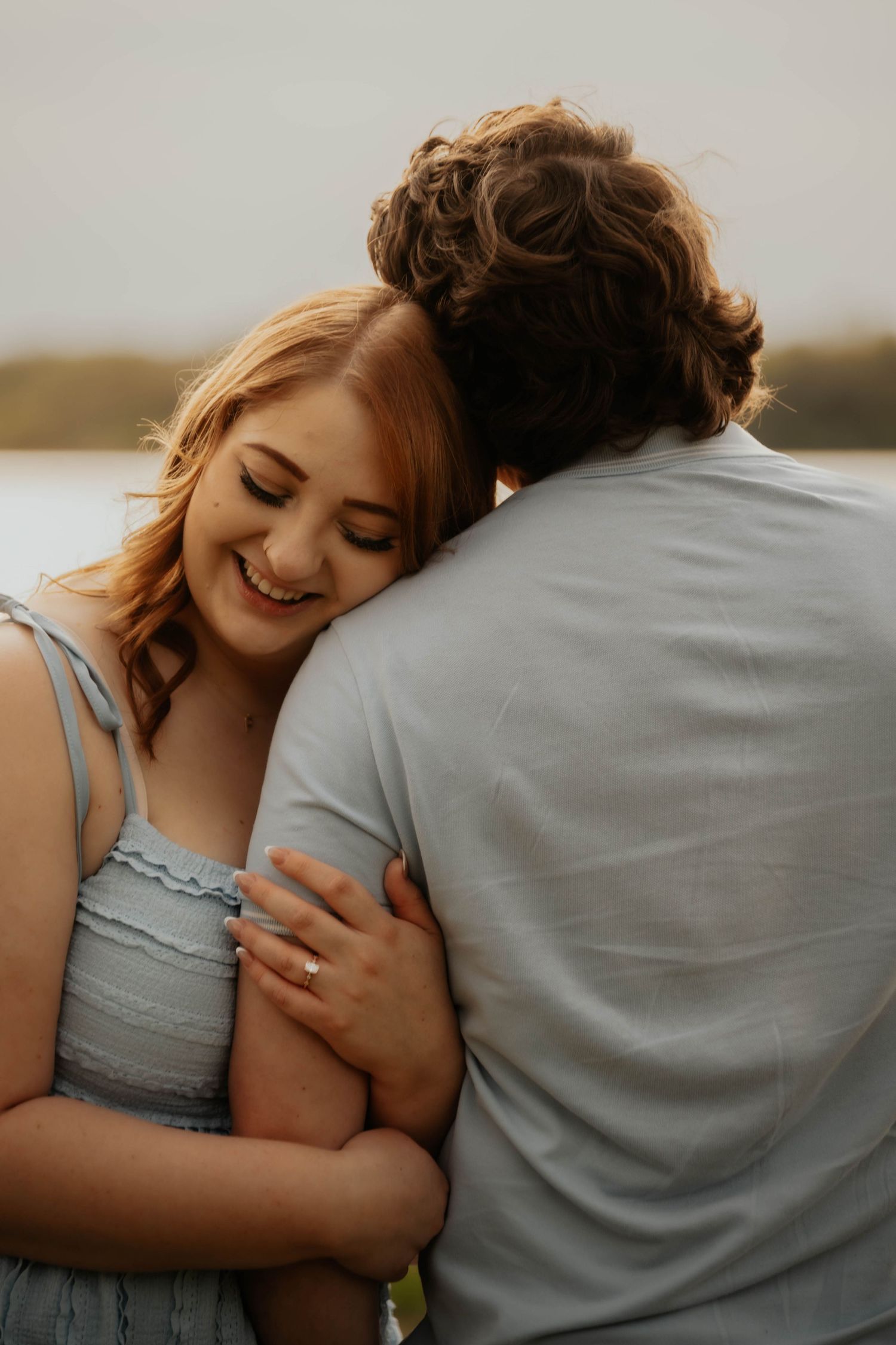 Smiling woman with engagement ring hugging her partner near a lake.