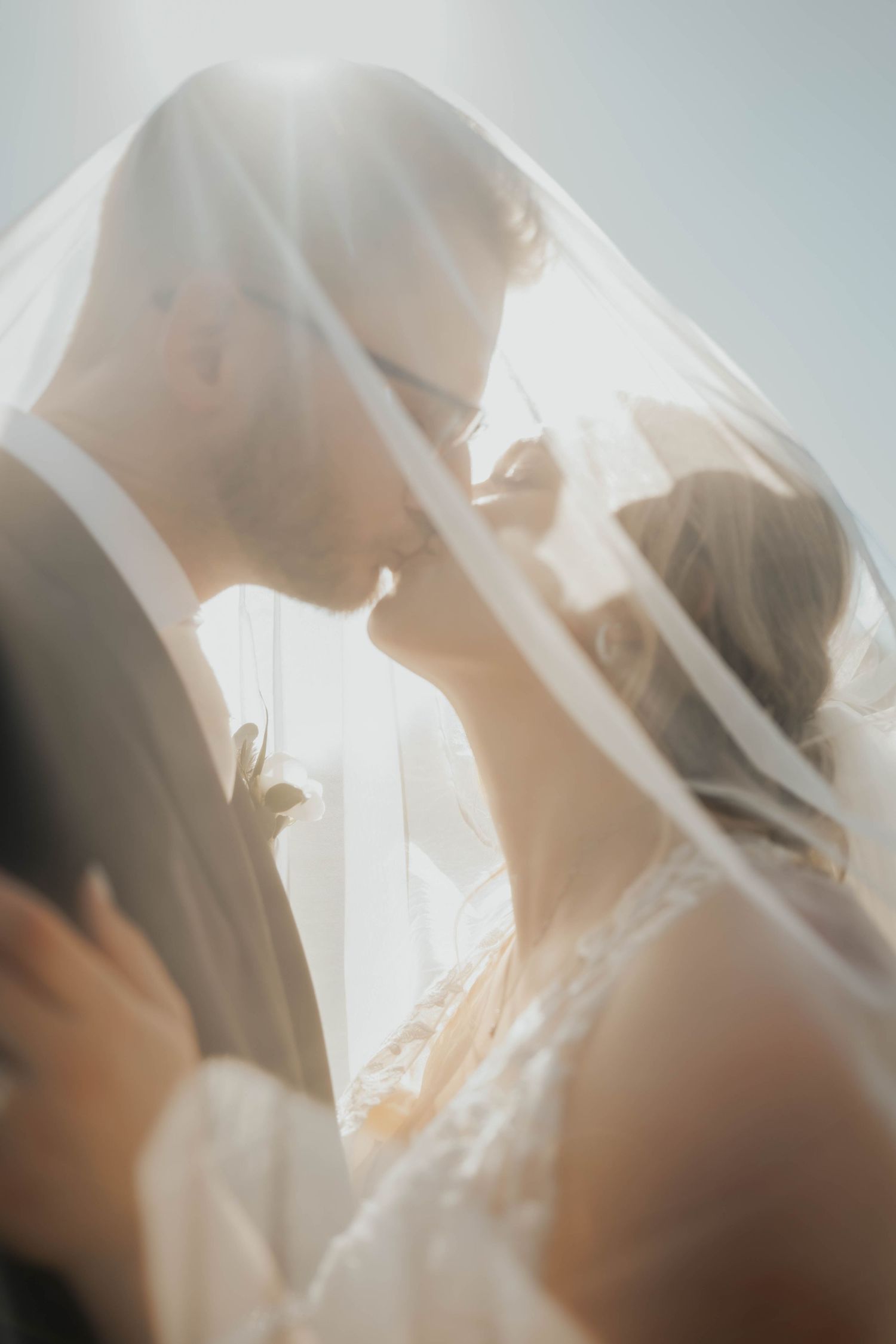 Bride and groom sharing a soft kiss under a flowing veil in dreamy backlit sunlight.