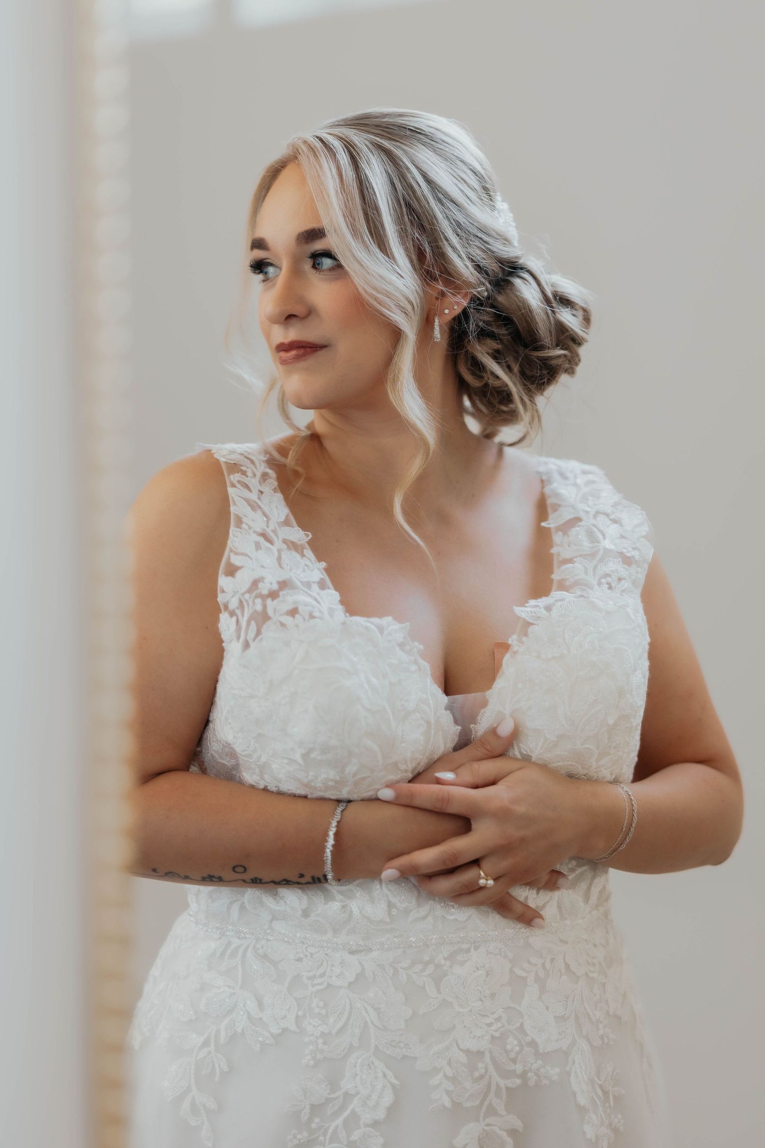 Elegant bride in lace wedding gown with updo hairstyle gazing thoughtfully near a bright window.