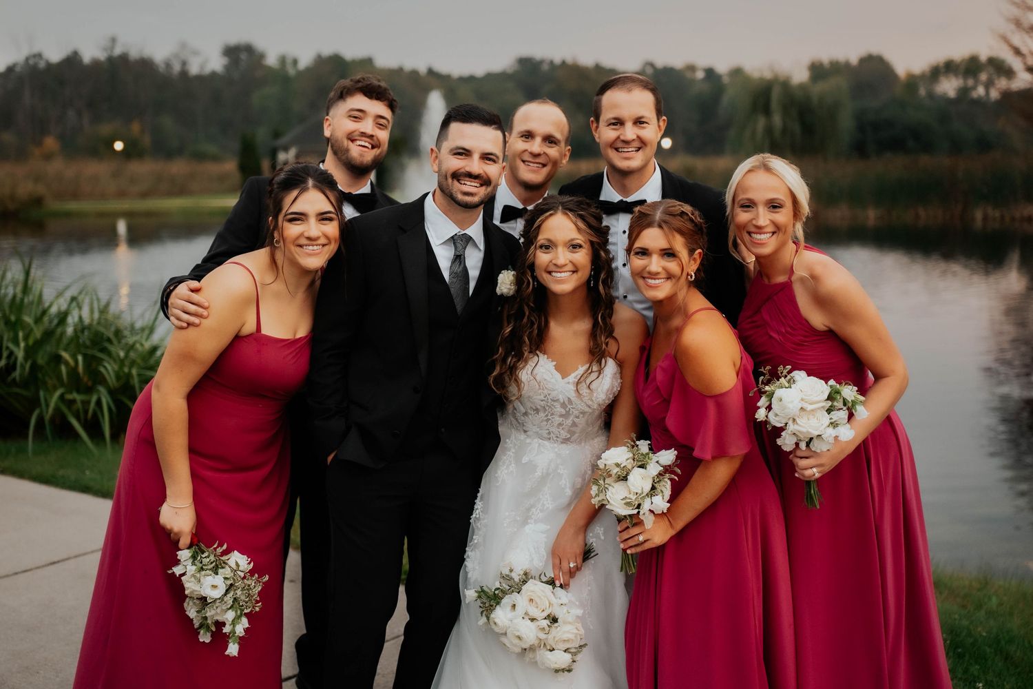 Wedding party in burgundy dresses and black suits posing together near a pond at outdoor reception.