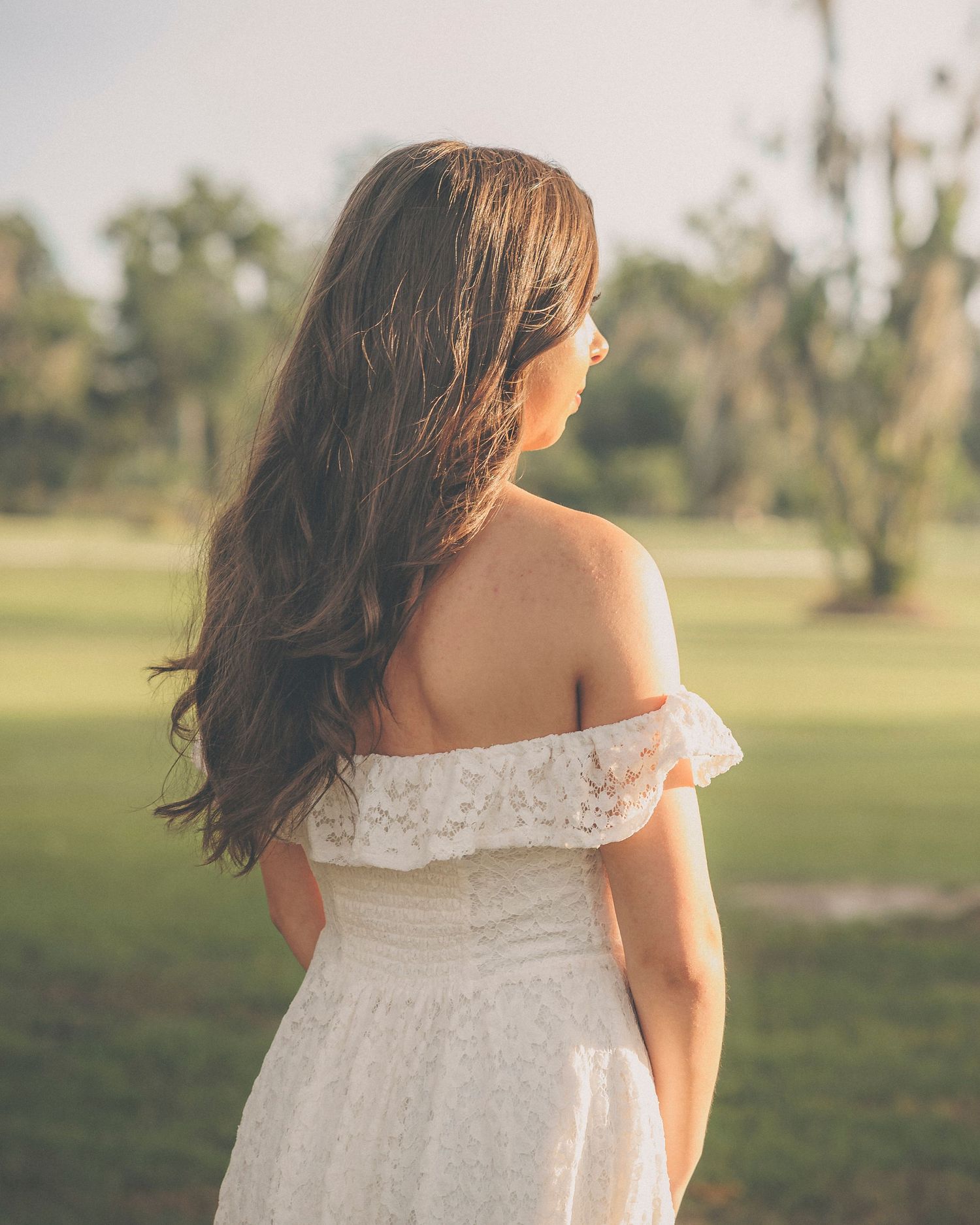 Candid outdoor photography in a white lace dress against a grassy field backdrop.