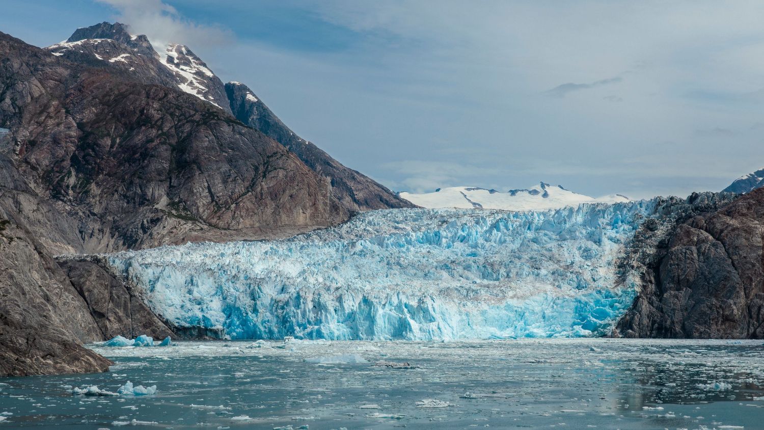 Massive blue glacier face meets the ocean in a dramatic Alaskan landscape panorama.