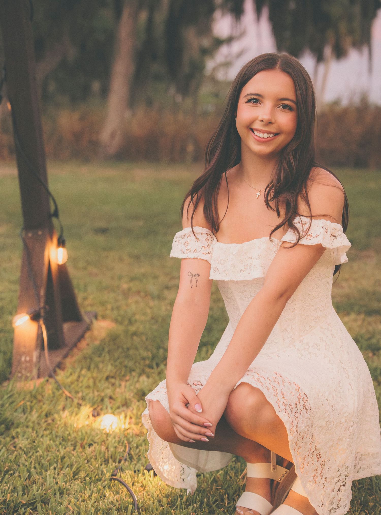 Graduate poses in white lace dress under outdoor string lights at sunset.