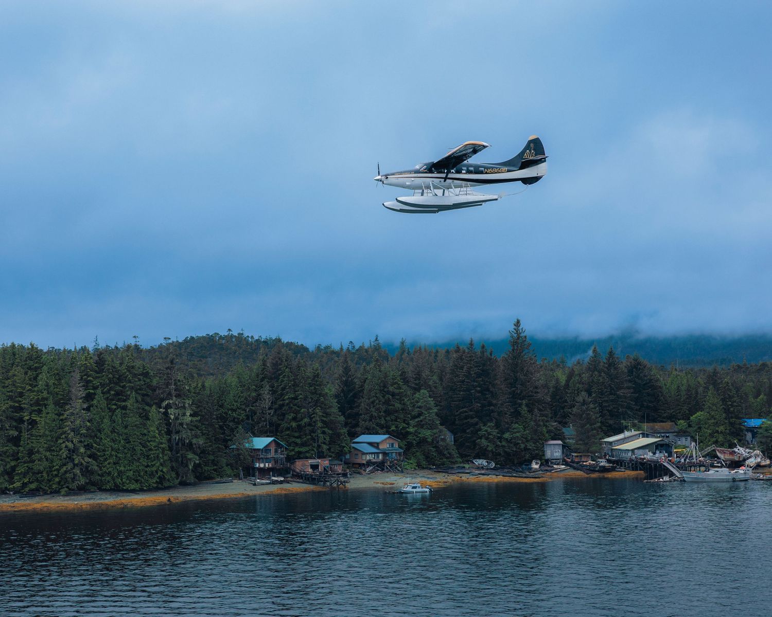 Floatplane flies over an Alaskan waterfront community nestled among evergreen forests.